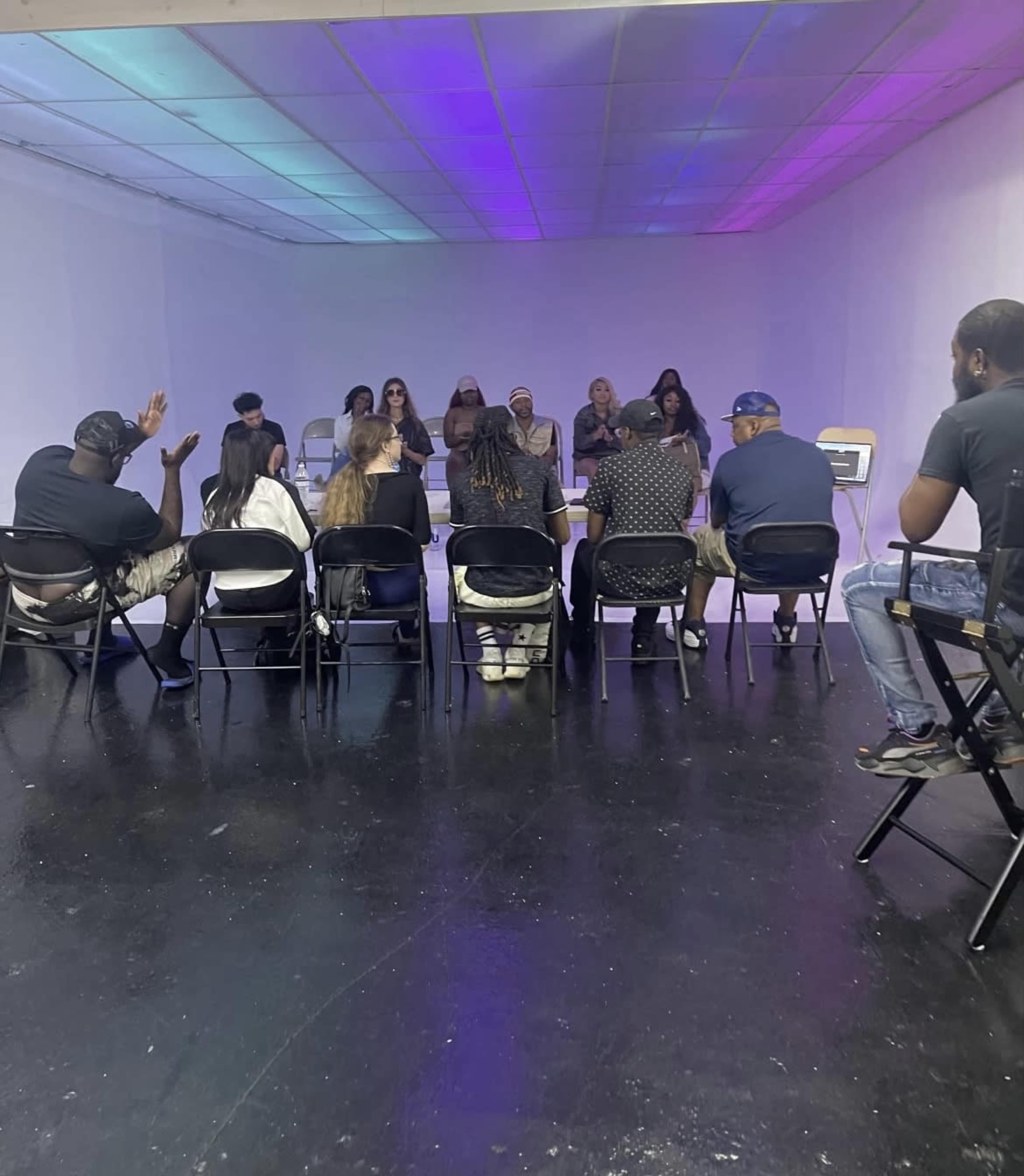 A group of people is seated in chairs facing a panel of individuals at a meeting in a dimly lit room.