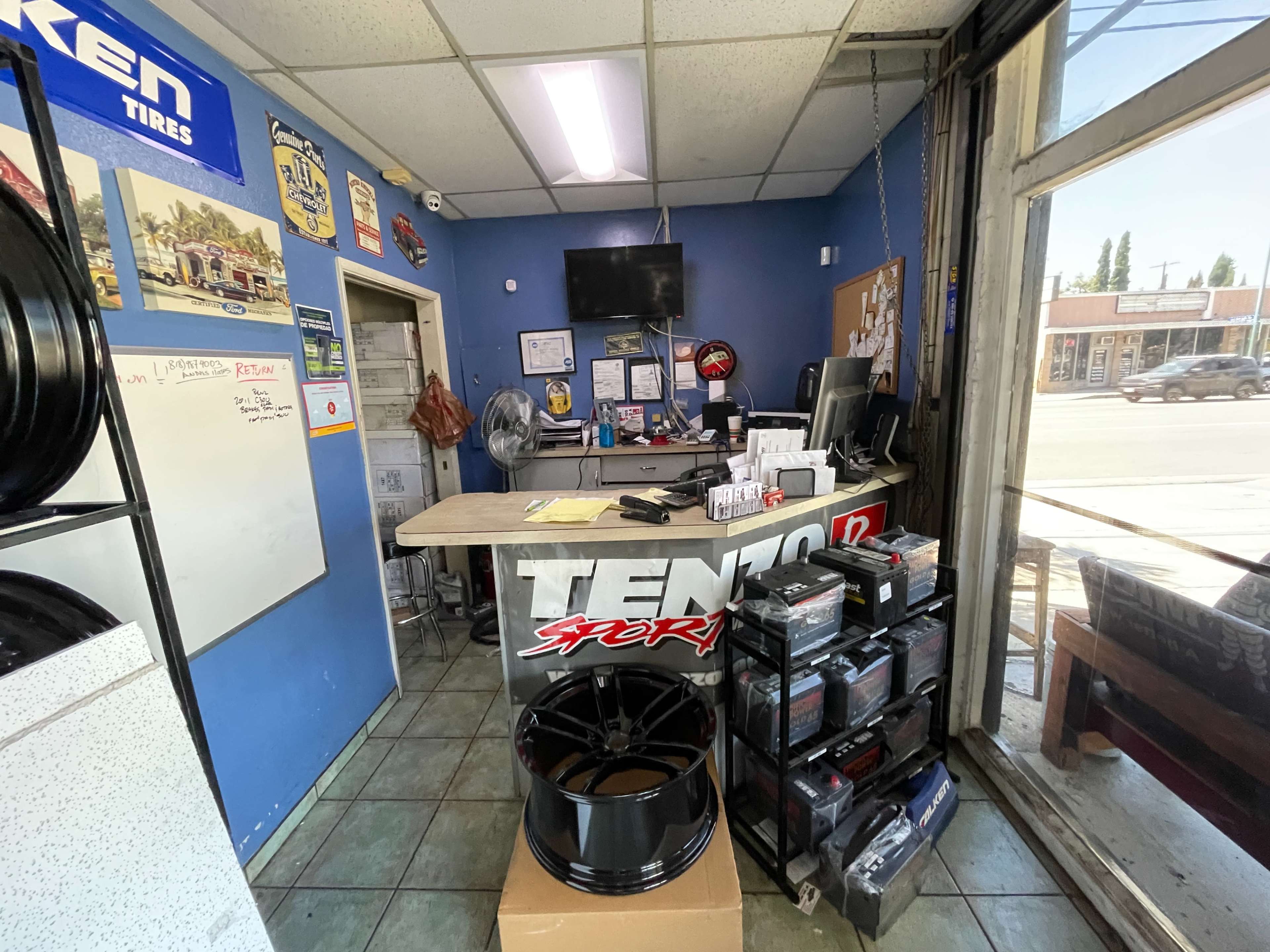 The image shows the interior of a tire shop with a counter displaying performance wheels and a rack of batteries, along with a fan and wall-mounted TV.