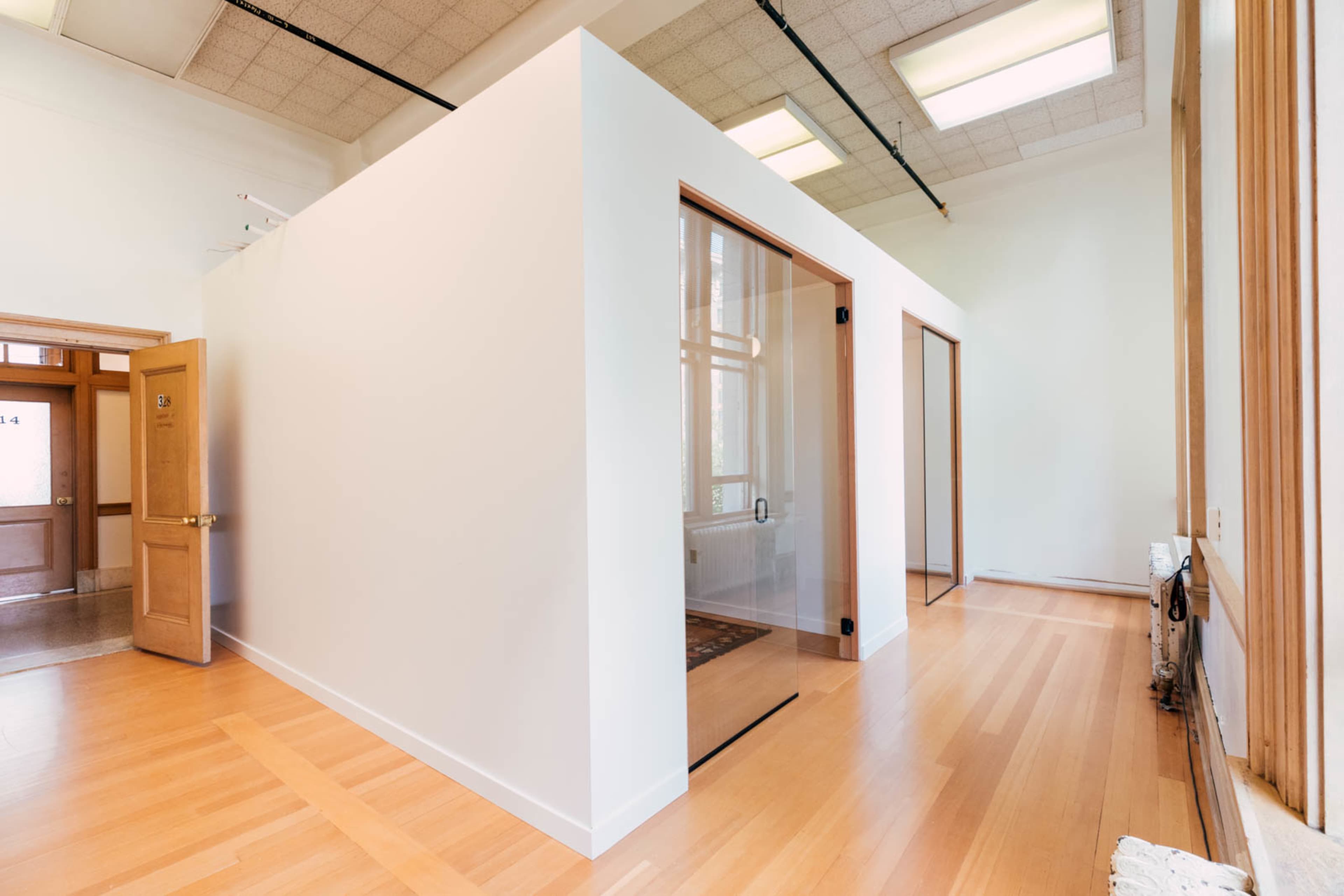 The image shows a bright, modern room with a white interior featuring light wood flooring and a glass-walled office space.