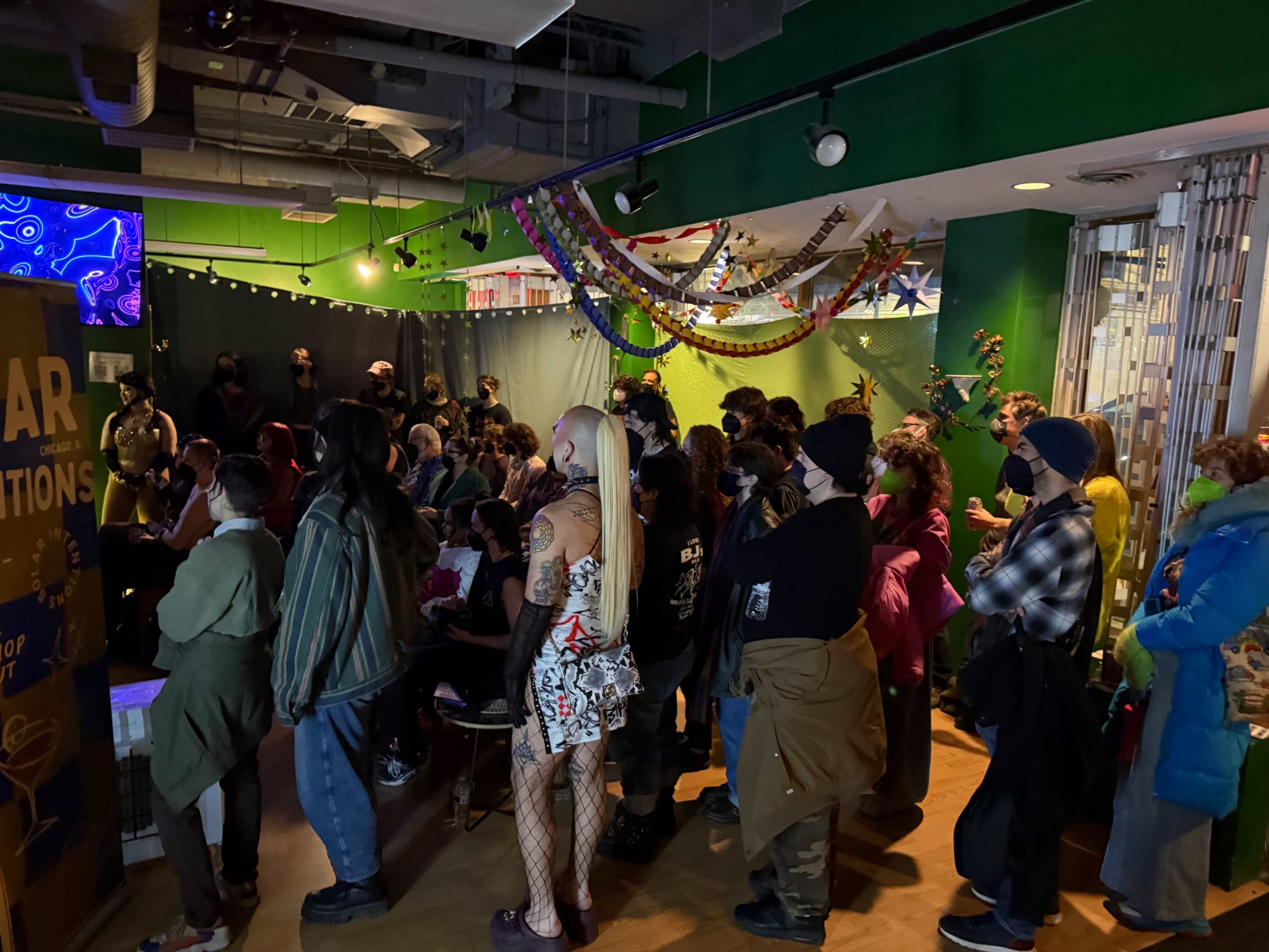 A crowd of people stands and sits in a dimly lit room, engaged in a presentation or performance, with colorful decorations hanging above.