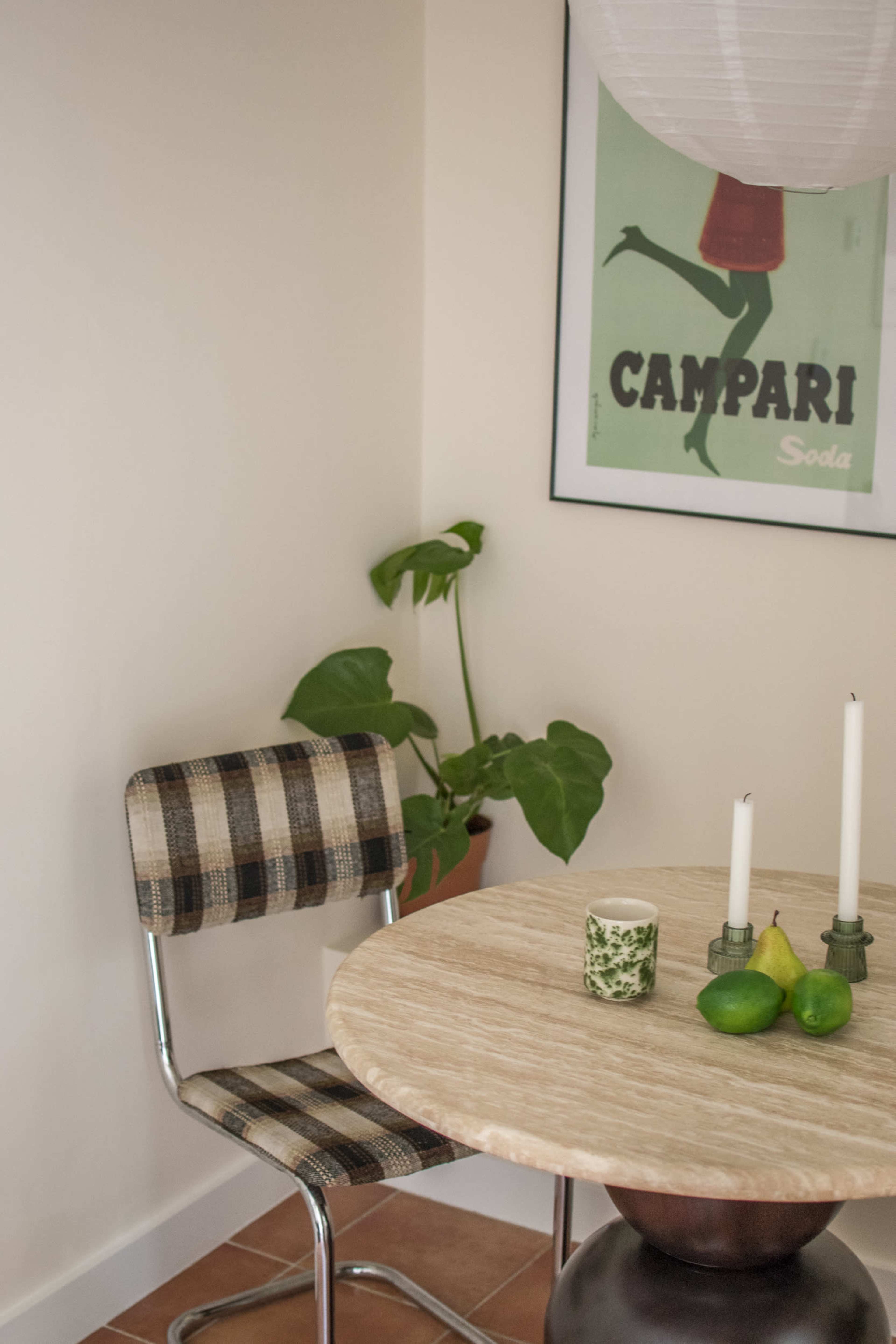 A round table with a marble top is set with two candles and a small arrangement of two limes and a pear, with a plaid chair nearby and a plant in the corner.