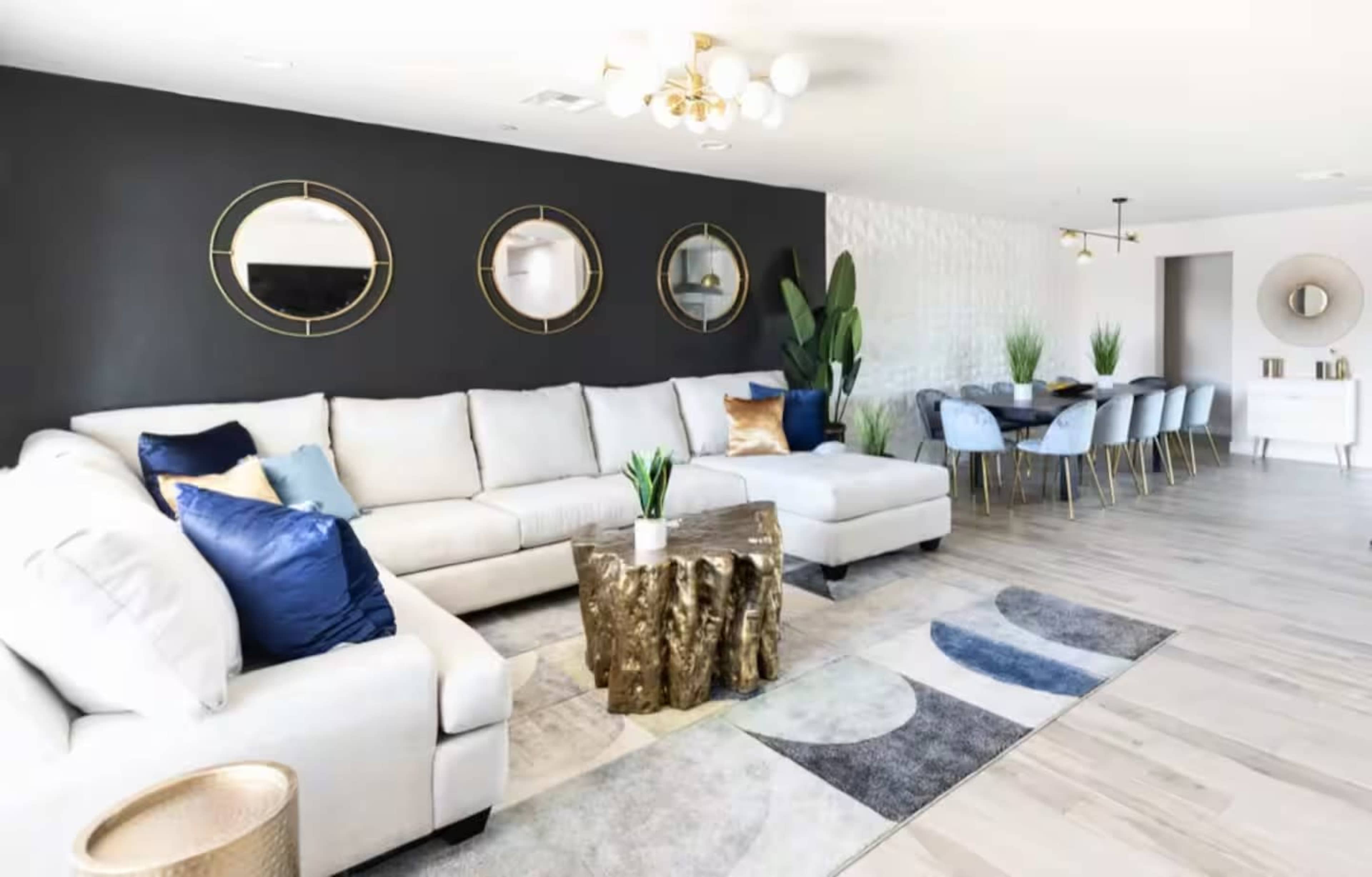 A modern living room features a large light-colored sectional sofa, three round mirrors on a dark wall, a wooden stump table, and a dining area with a table and chairs.