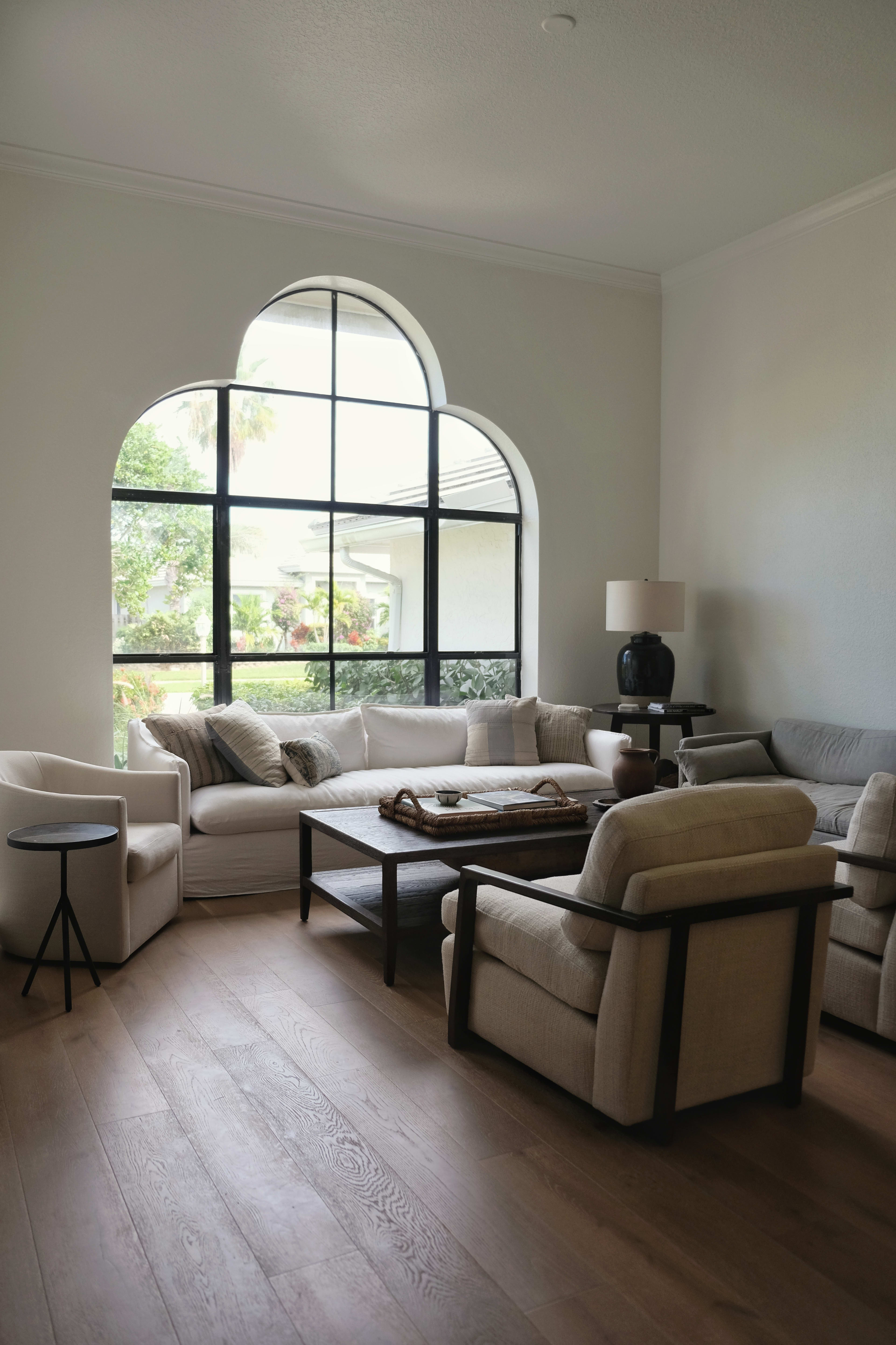 The image shows a living room with a large arched window, furnished with a mix of armchairs and sofas around a coffee table on wooden flooring.