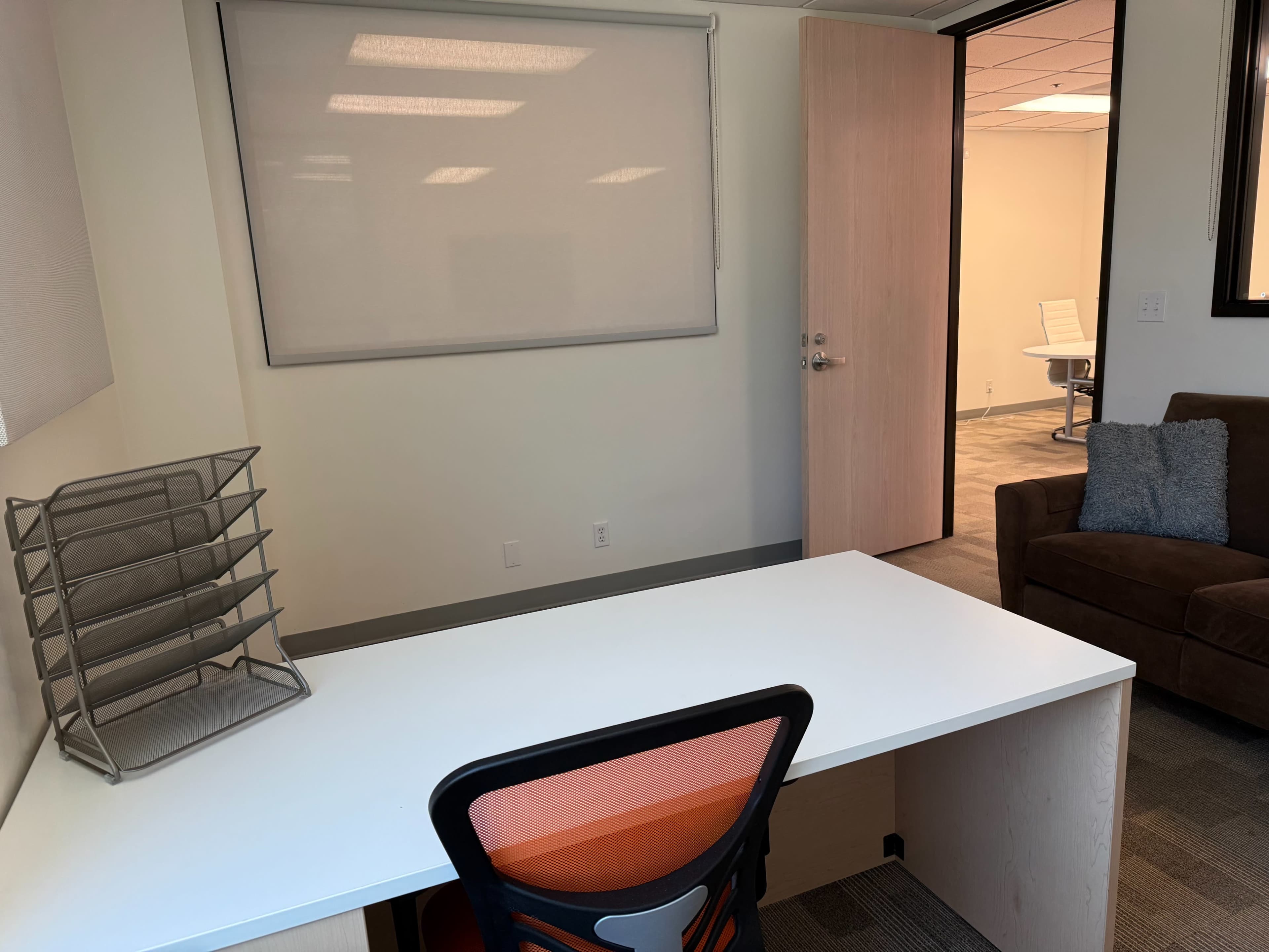 The image shows an office space featuring a white desk with an orange office chair, a file organizer, and a brown couch, along with an open doorway leading to another room.