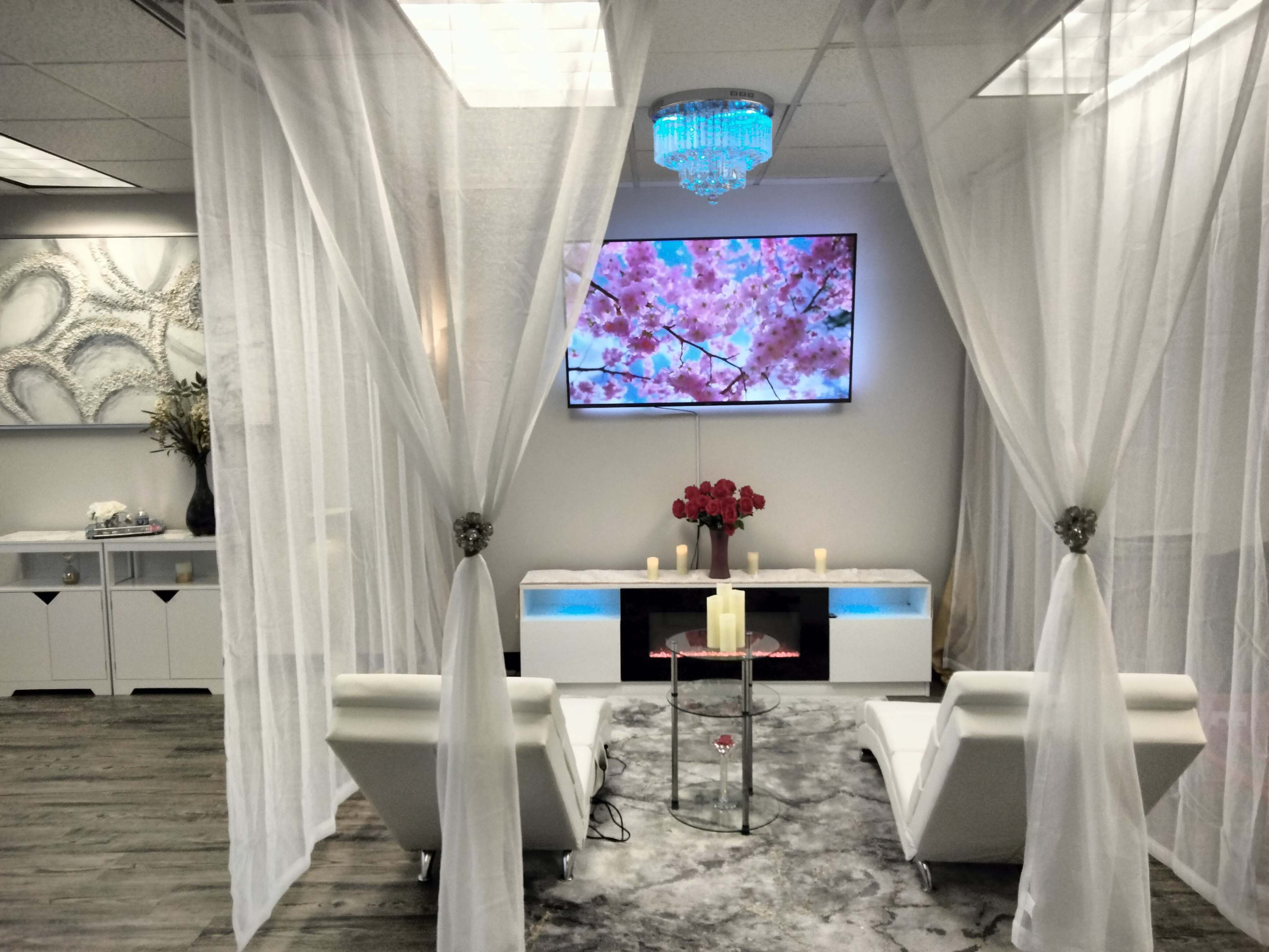 A serene lounge area featuring two white chairs, a glass table, and a large television displaying floral imagery, all surrounded by sheer white curtains.