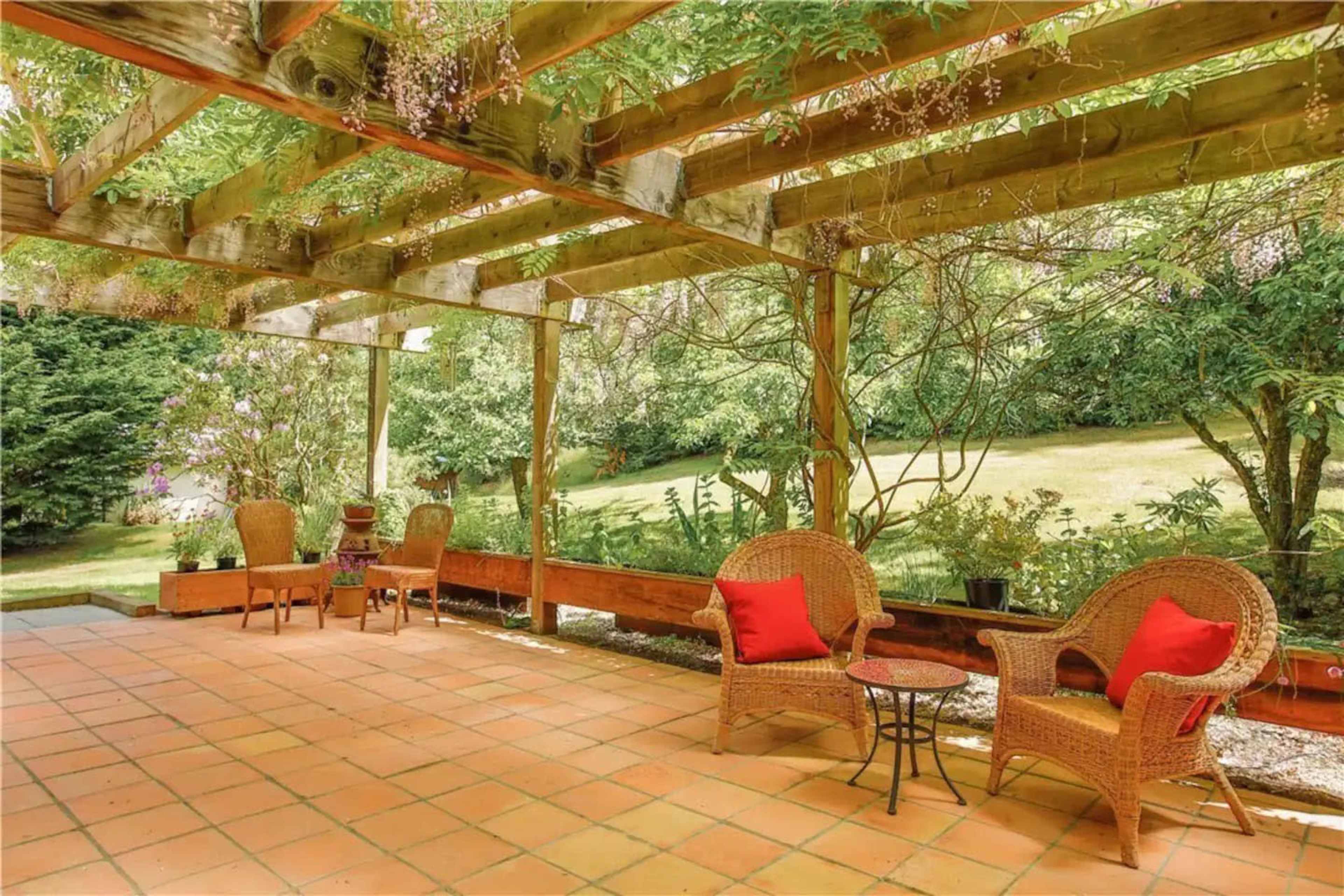 The image shows a patio area with wicker furniture and red cushions under a wooden pergola adorned with greenery.
