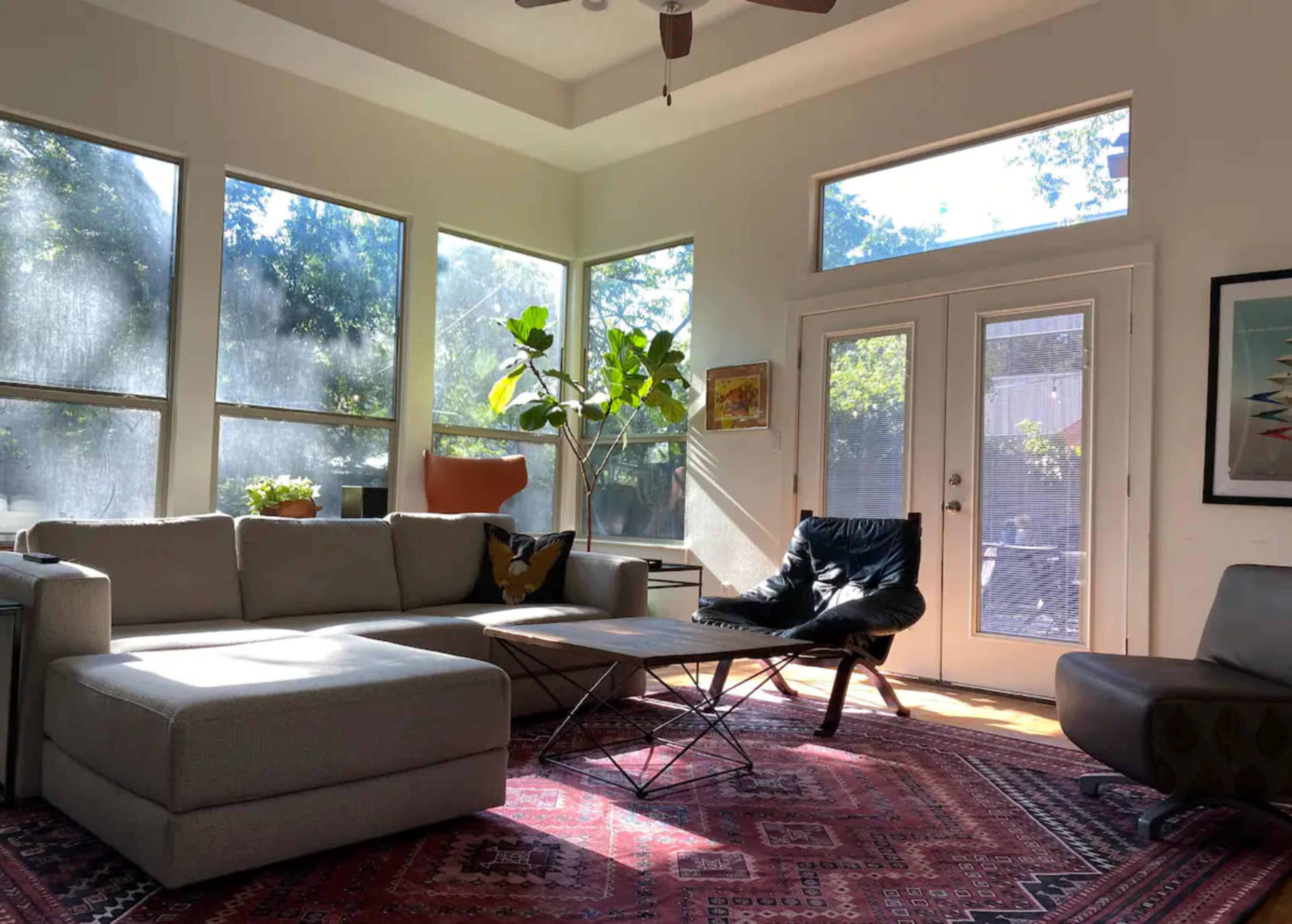 A well-lit living room with large windows, a grey sectional sofa, a black chair, and a coffee table on a patterned rug.