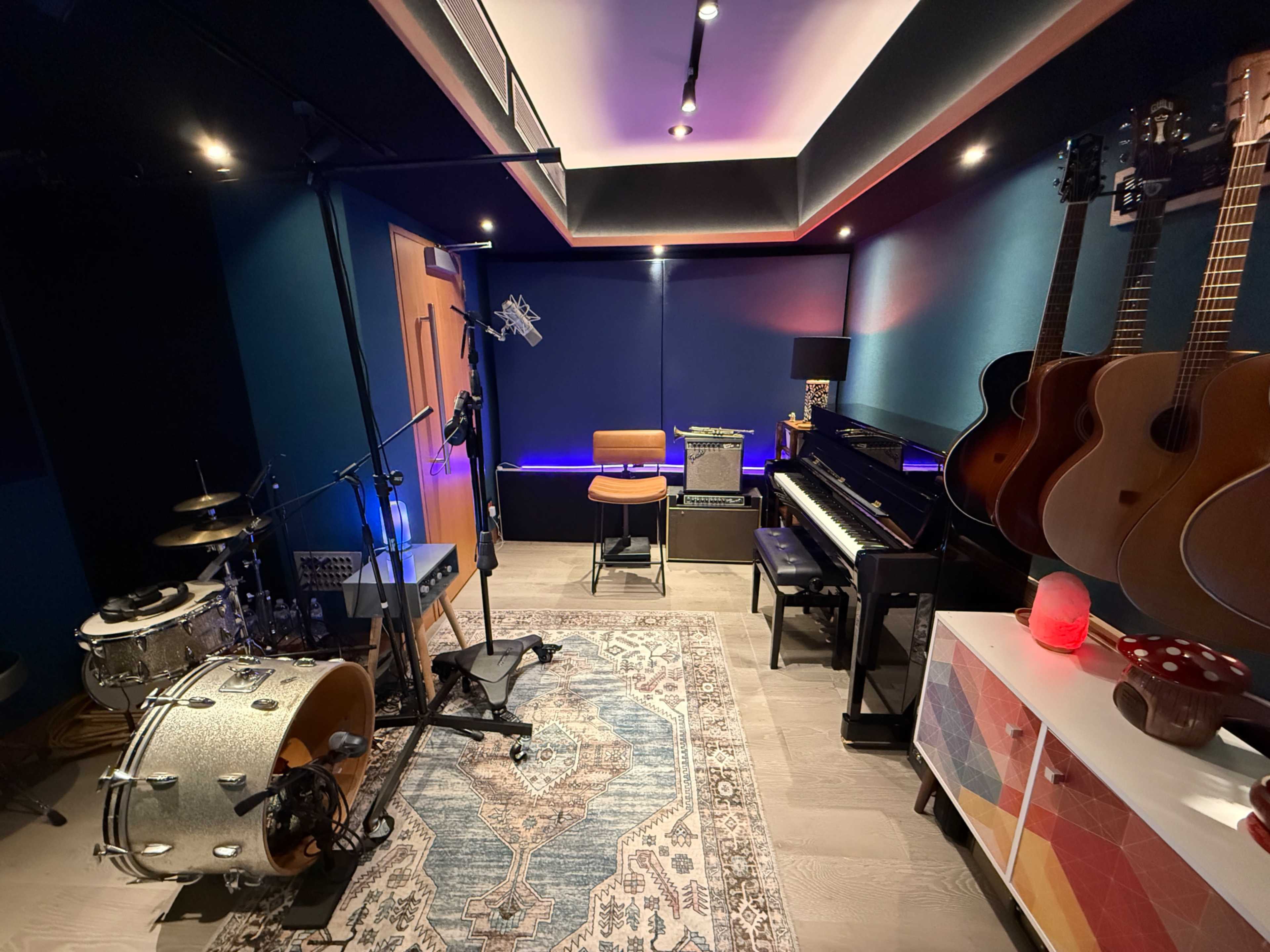 The image shows a small music studio featuring a drum set, a piano, several guitars mounted on the wall, and a colorful area rug.