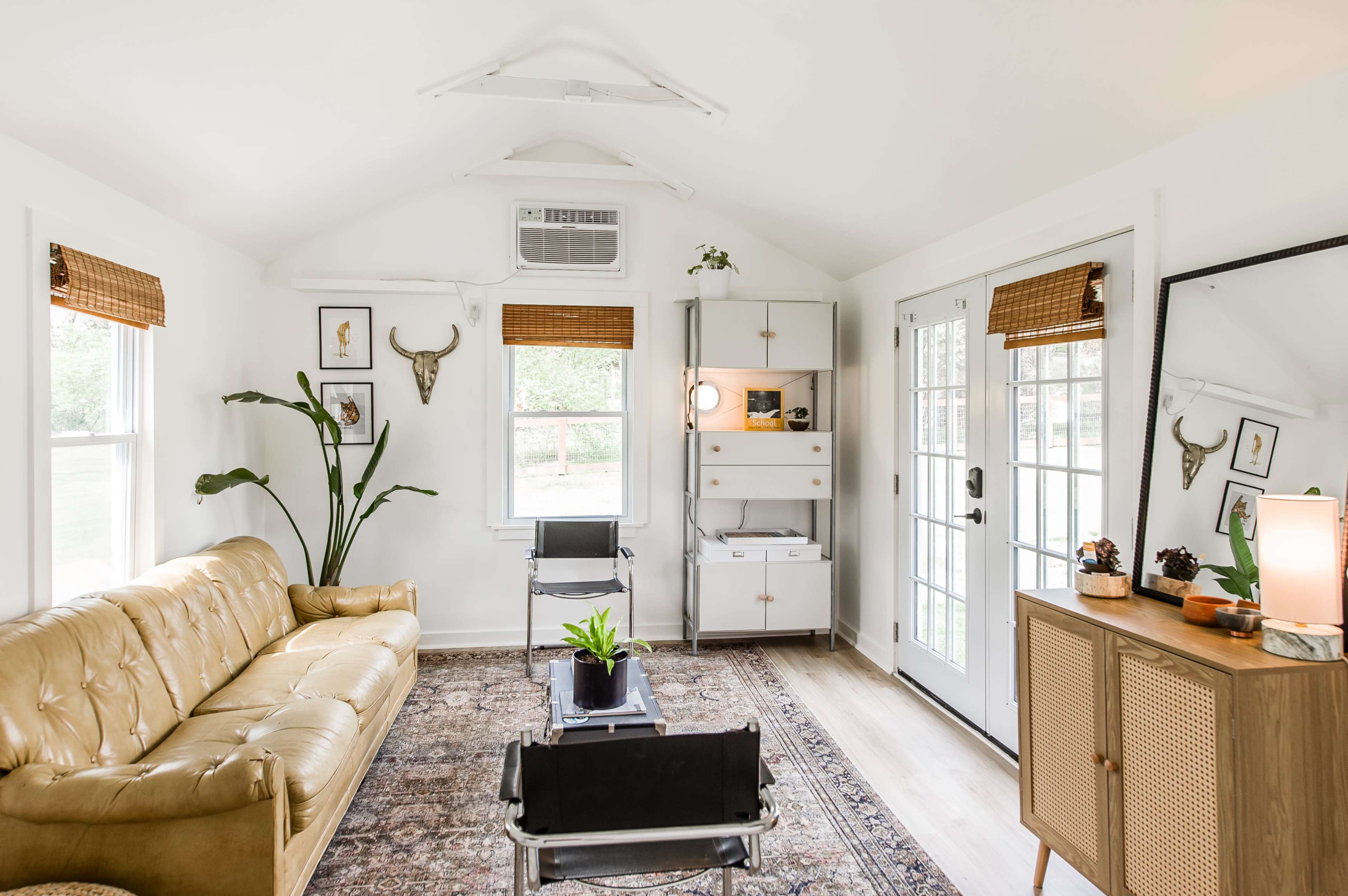 The image shows a bright, small living room featuring a tan leather sofa, a decorative rug, a wooden cabinet, and large windows with light filtering through.