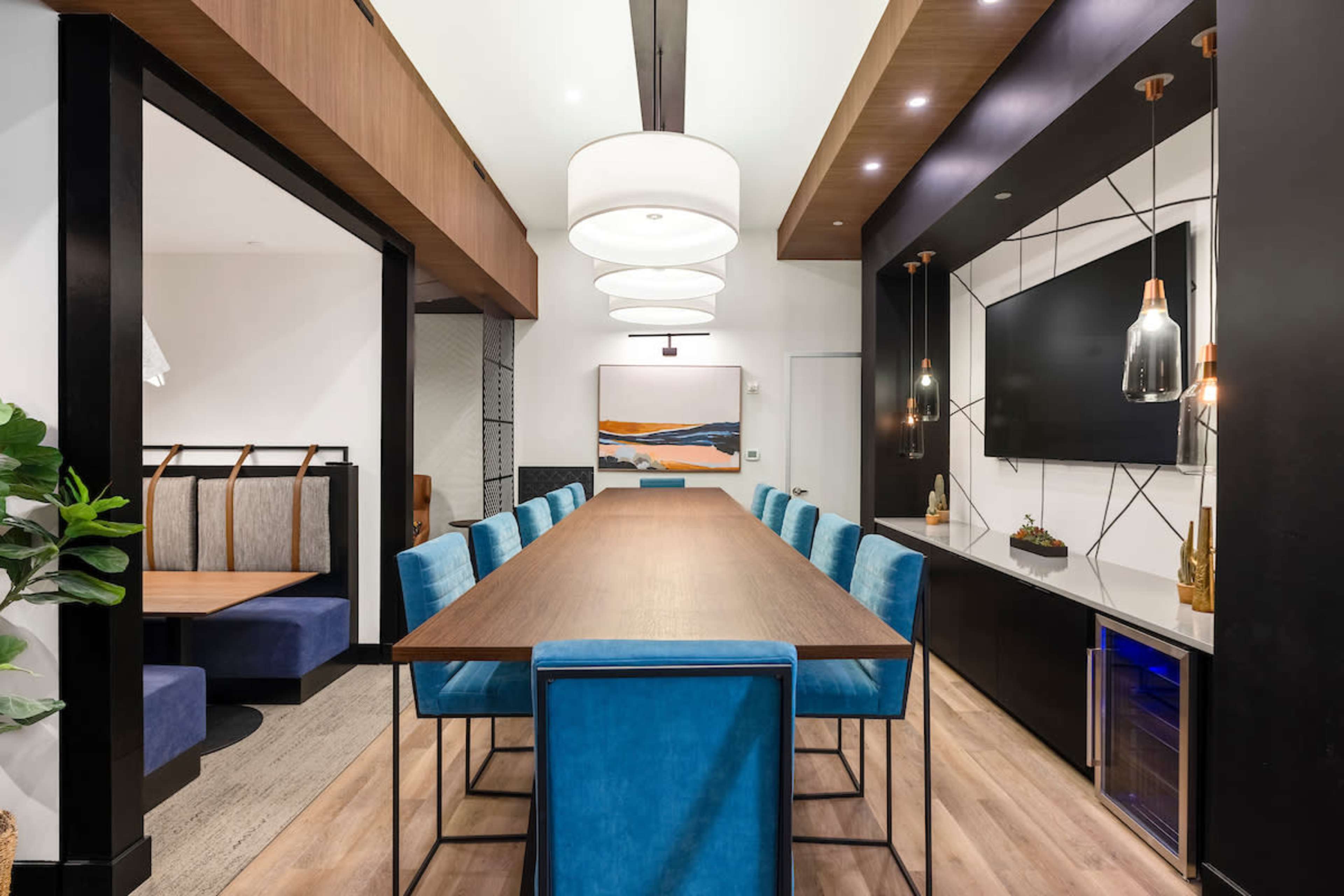 A dining area with a long wooden table surrounded by blue chairs, illuminated by modern pendant lights and featuring a large TV mounted on a black wall.