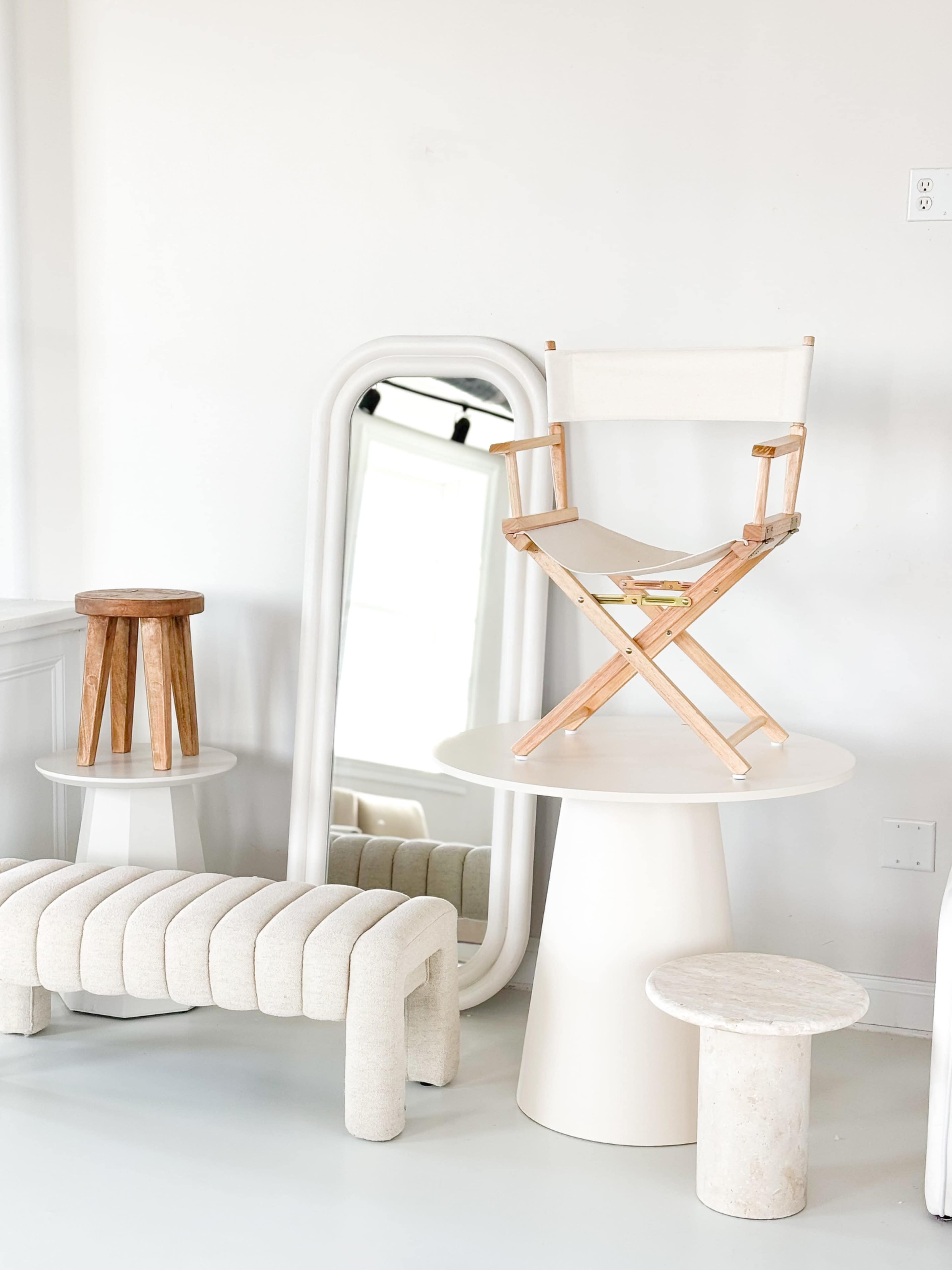 The image features a minimalist interior scene with a wooden director's chair placed on a round table, alongside a large mirror and various other furniture pieces.