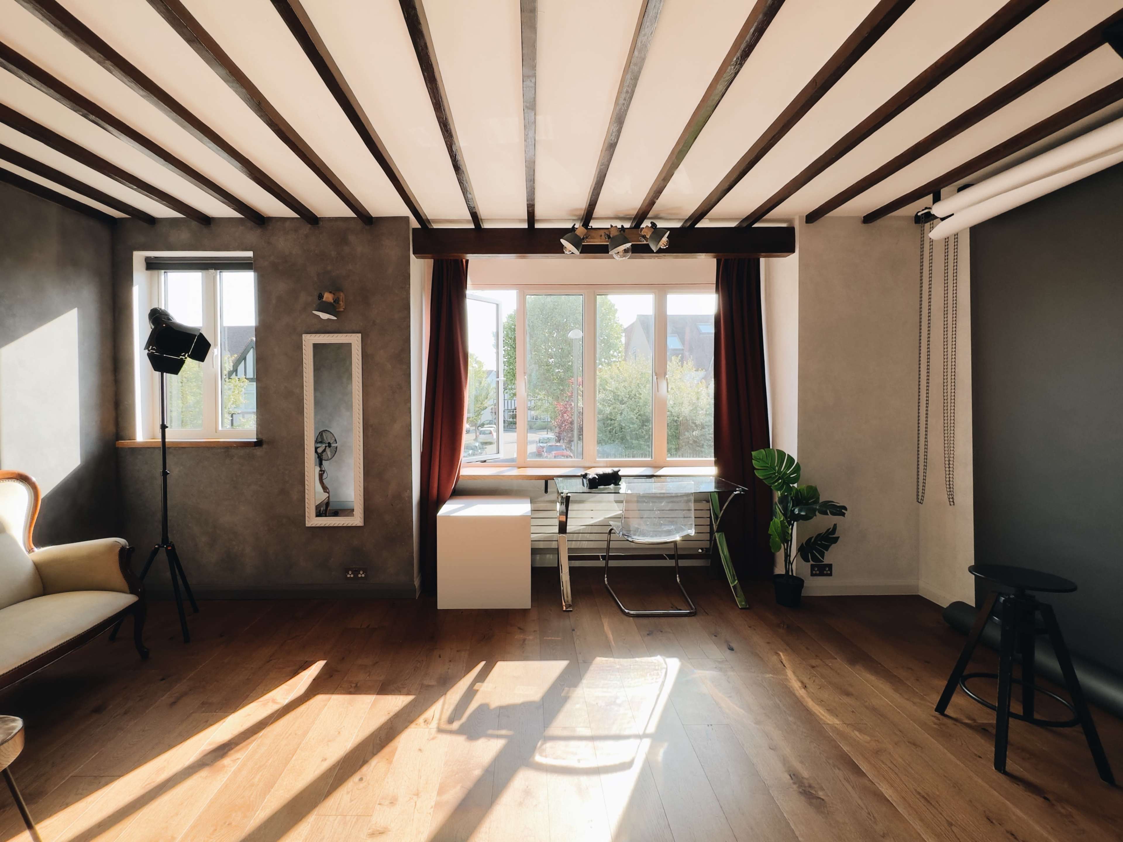 The room features wooden beams overhead, a large window with curtains, a light-colored sofa, a modern chair, and a potted plant near the corner.