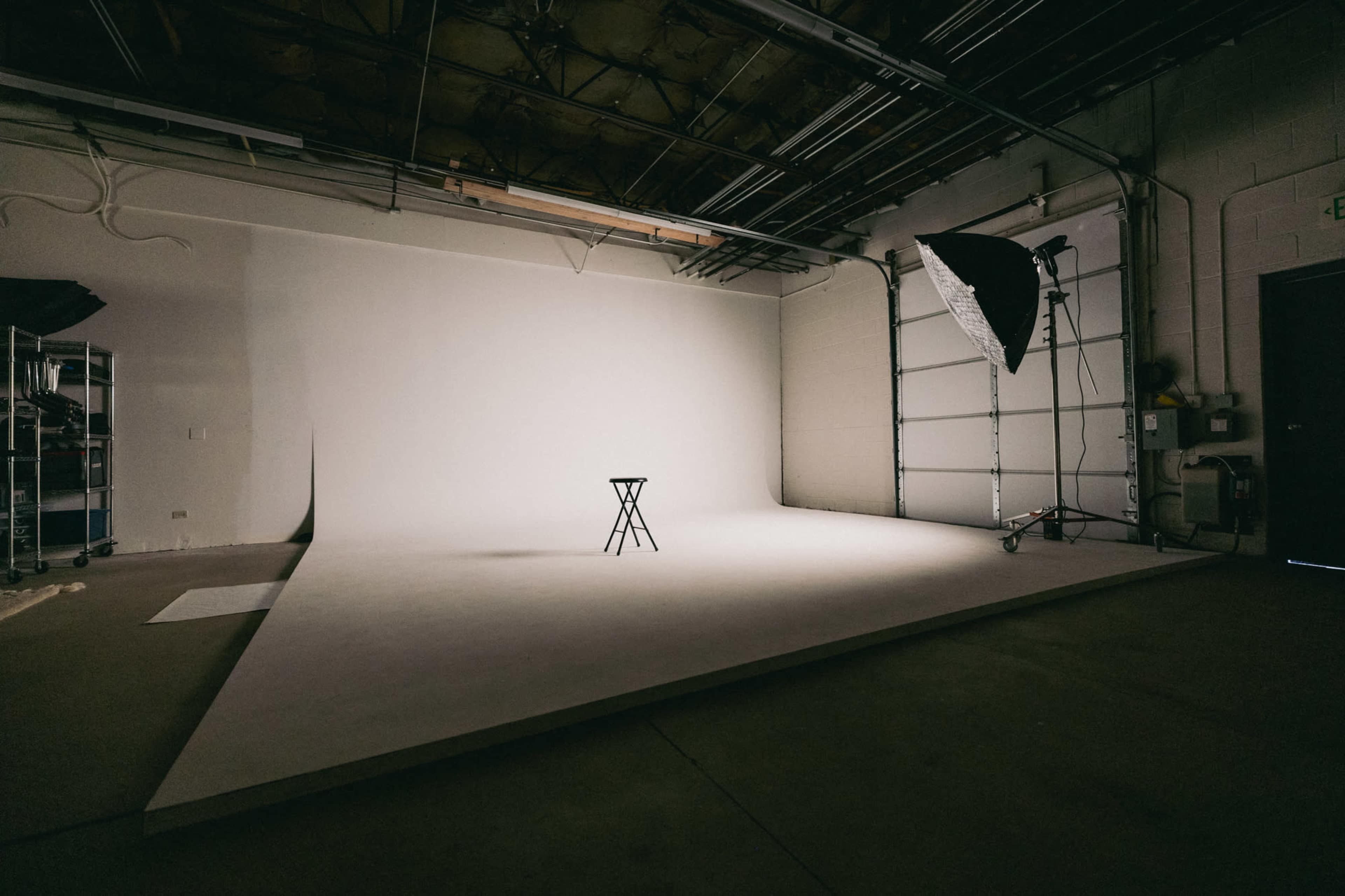 A minimalist photography studio features a single stool positioned in front of a blank backdrop, illuminated by a soft light source.
