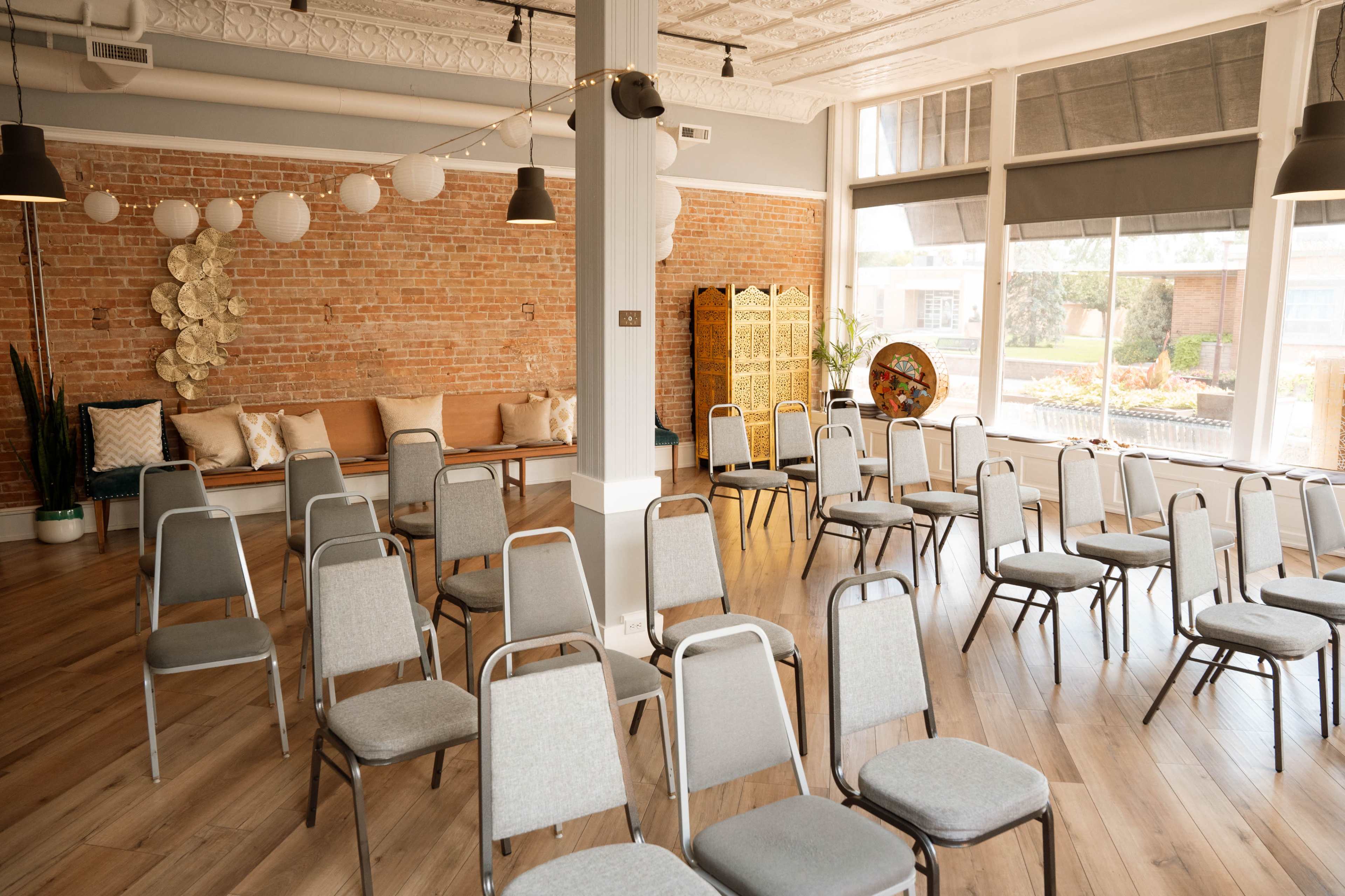 The image shows a spacious room with rows of metal-framed chairs set up for an event, featuring exposed brick walls and a small seating area with decorative pillows.