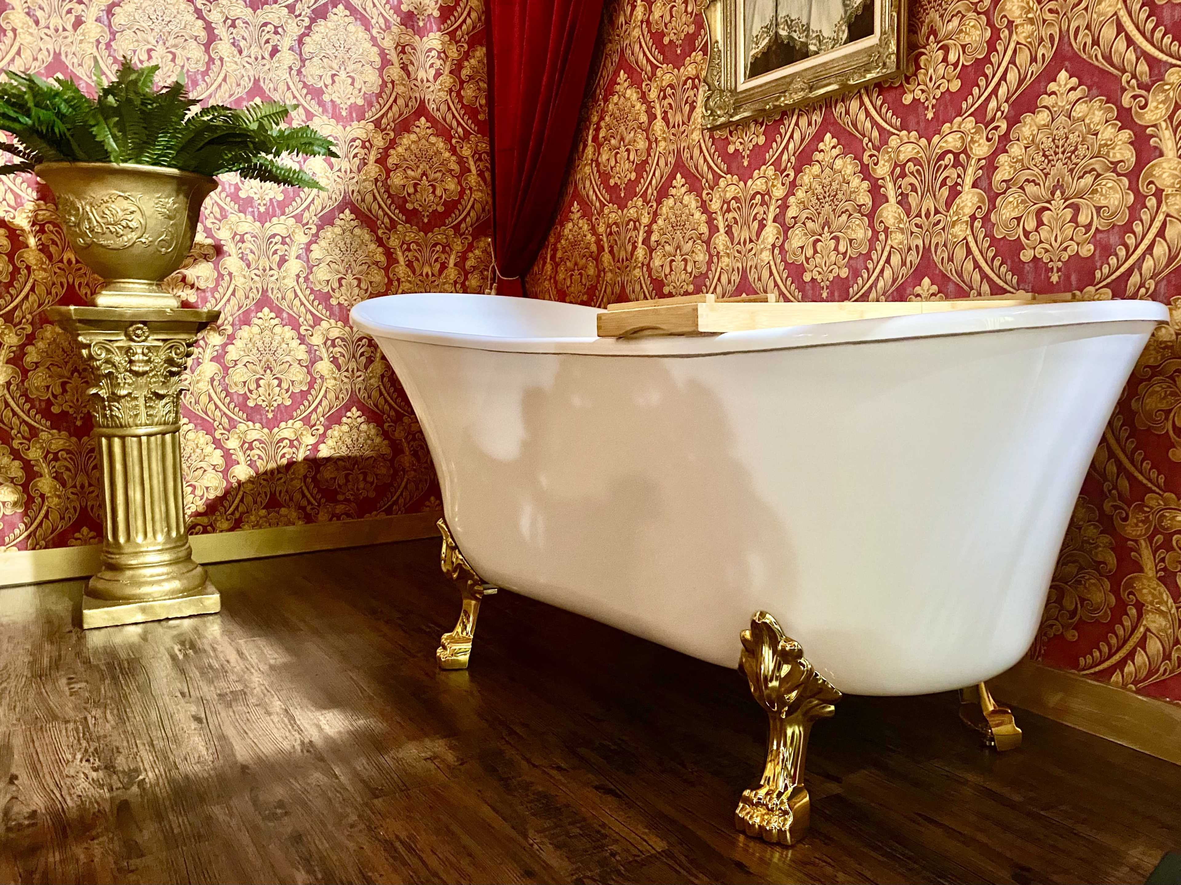 A white clawfoot bathtub with gold legs sits in a room decorated with ornate red wallpaper and a matching gold-accented plant stand.
