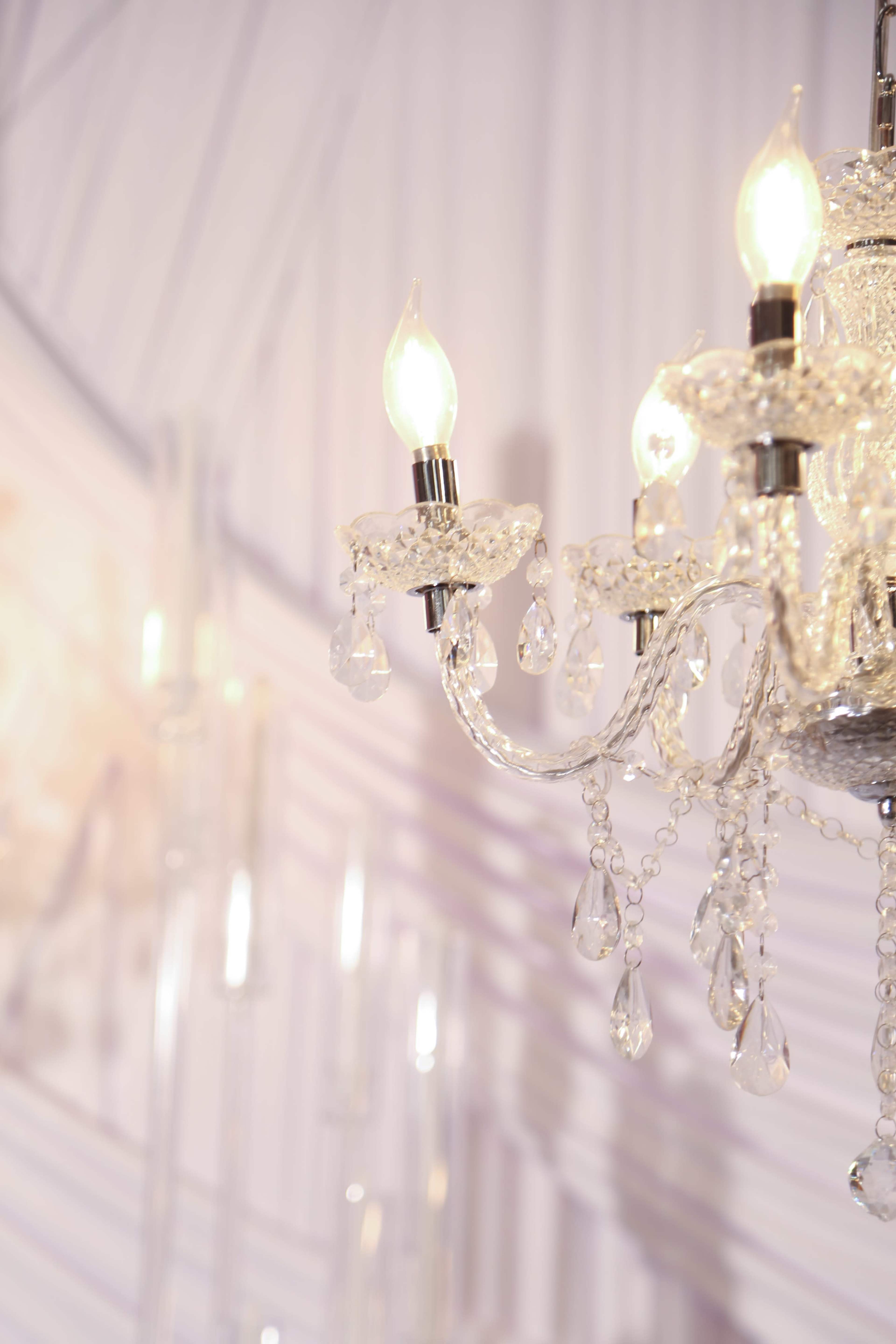 A crystal chandelier with three lit bulbs hangs against a backdrop of soft draped fabric.