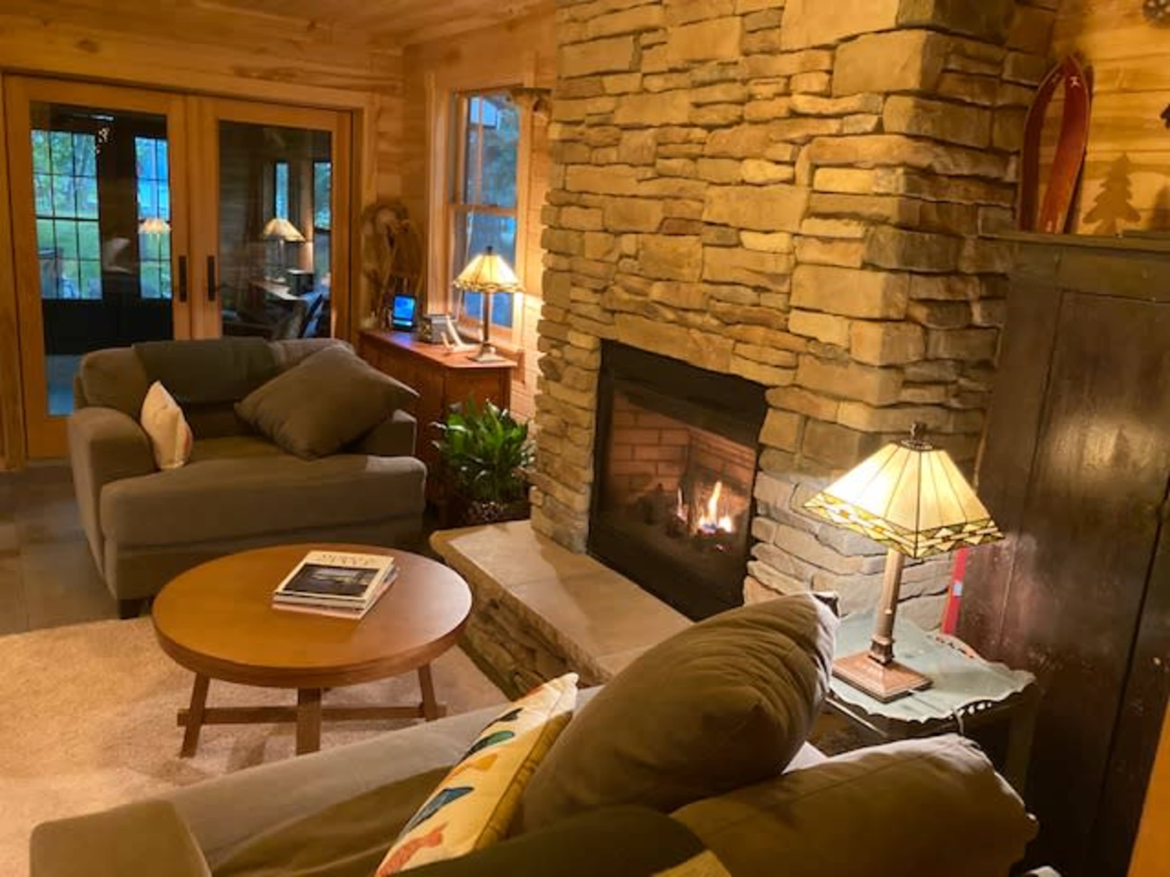 A cozy living room features a stone fireplace with a glowing fire, surrounded by comfortable seating and warm lighting.