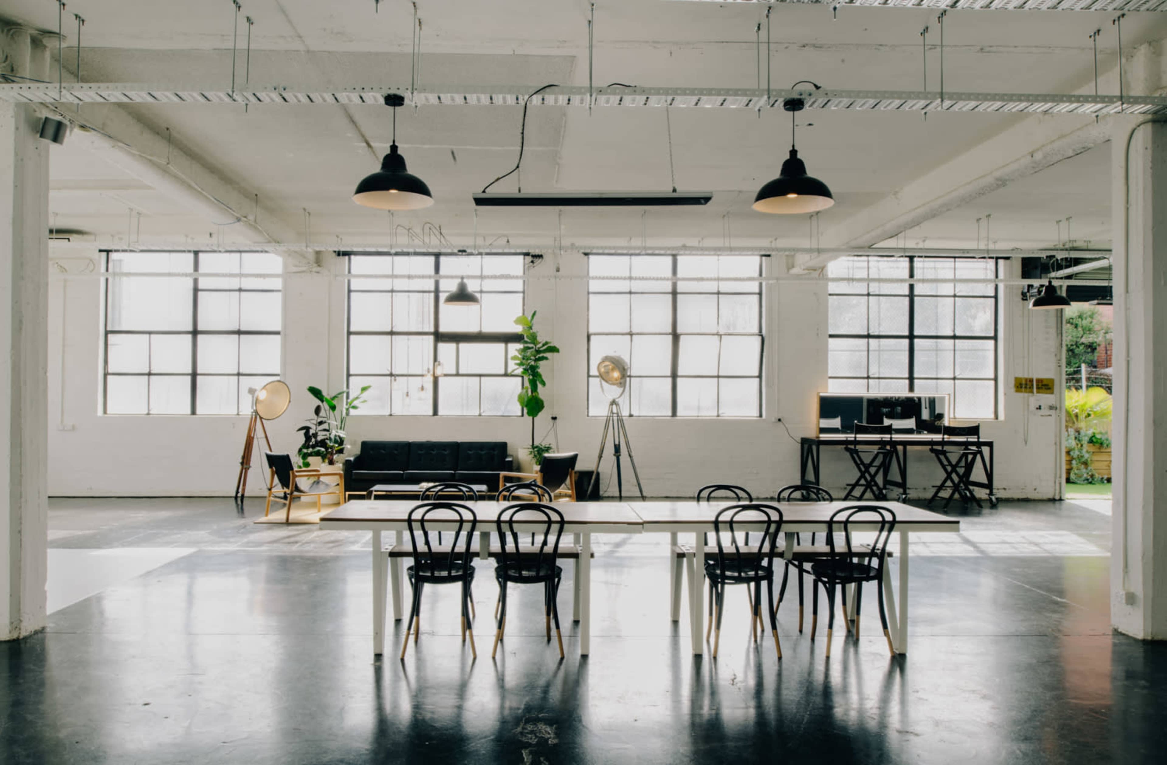The image shows a spacious, modern office with large windows, a seating area, a long table surrounded by chairs, and decorative plants.