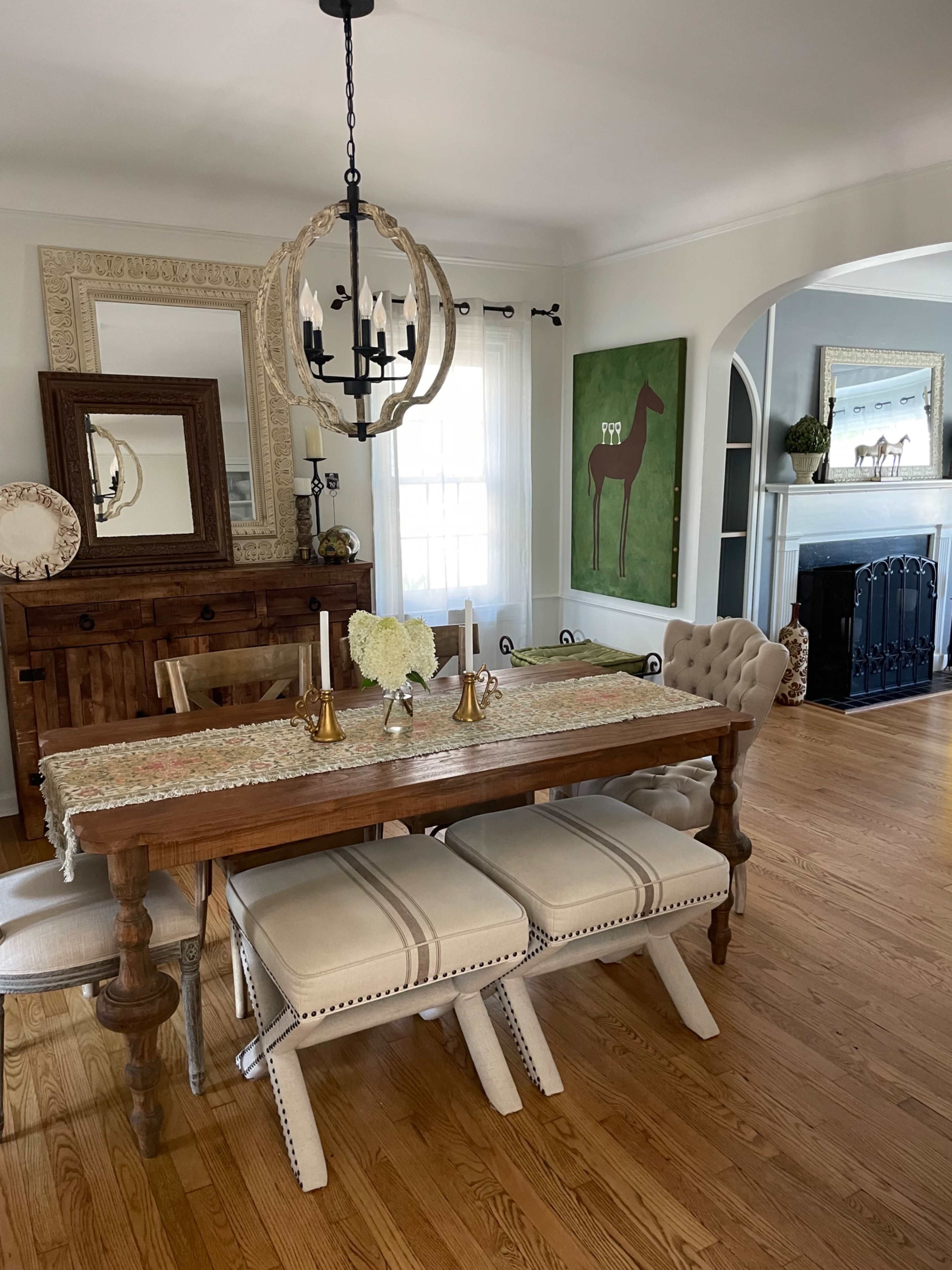 A wooden dining table with a runner and candles is set in a bright room featuring a chandelier, beige chairs, and decorative artwork on the walls.