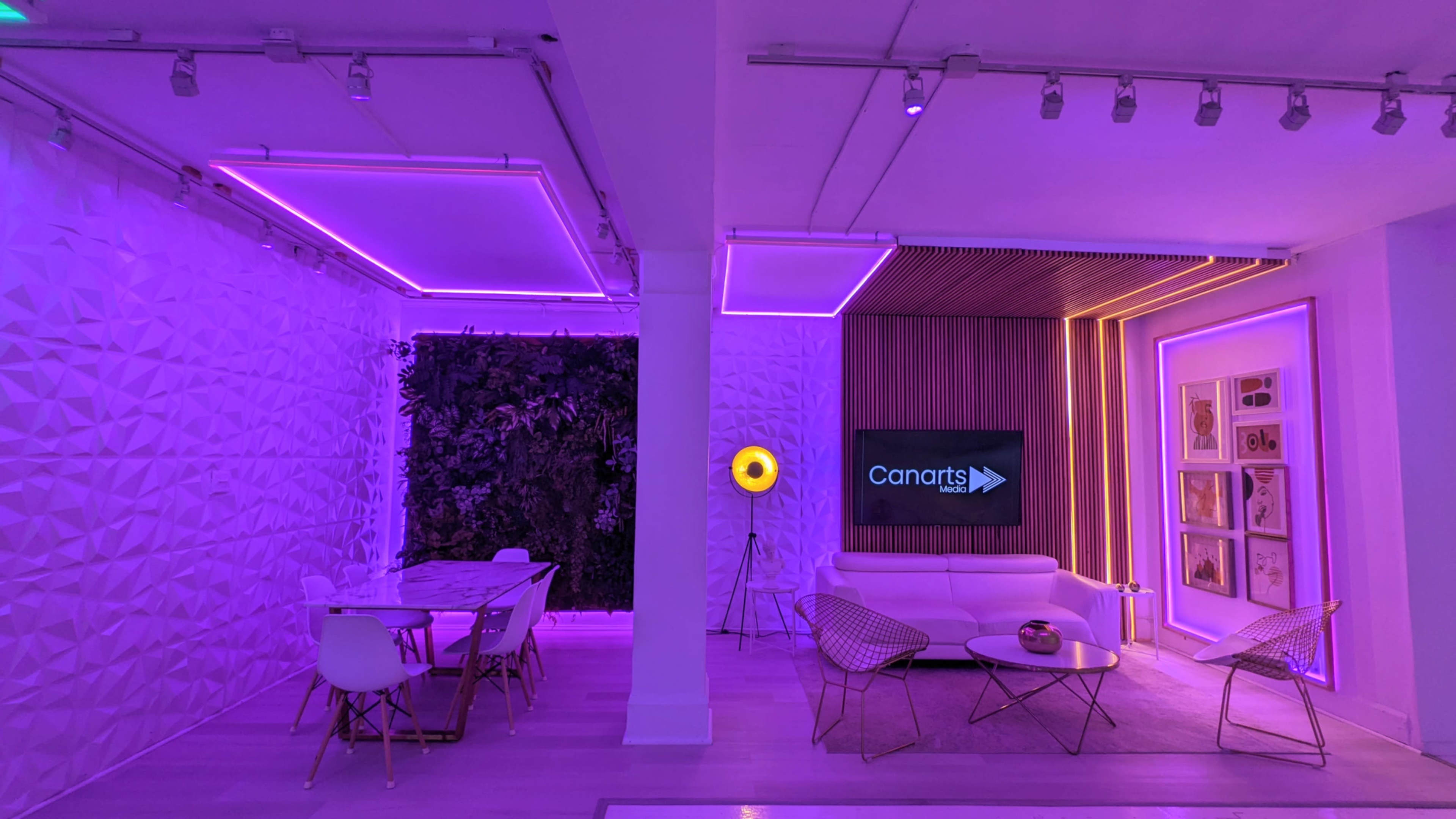 A modern interior space featuring a lounge area with a white sofa, a dining table, and ambient purple lighting, along with greenery and a wall-mounted screen displaying the logo "Canarts."