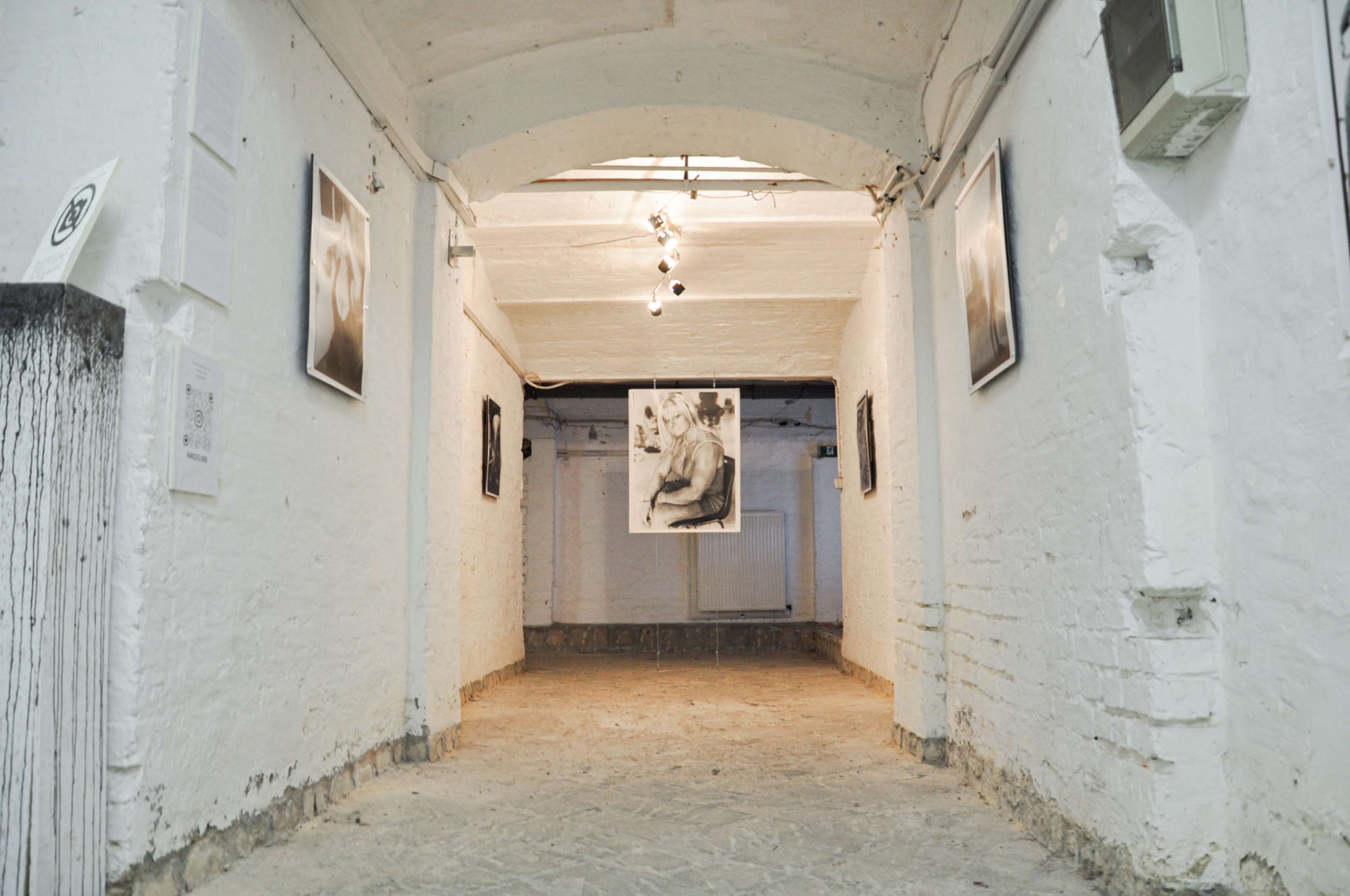 The image shows a narrow, white-walled corridor in an exhibition space, featuring black-and-white photographs displayed on the walls.
