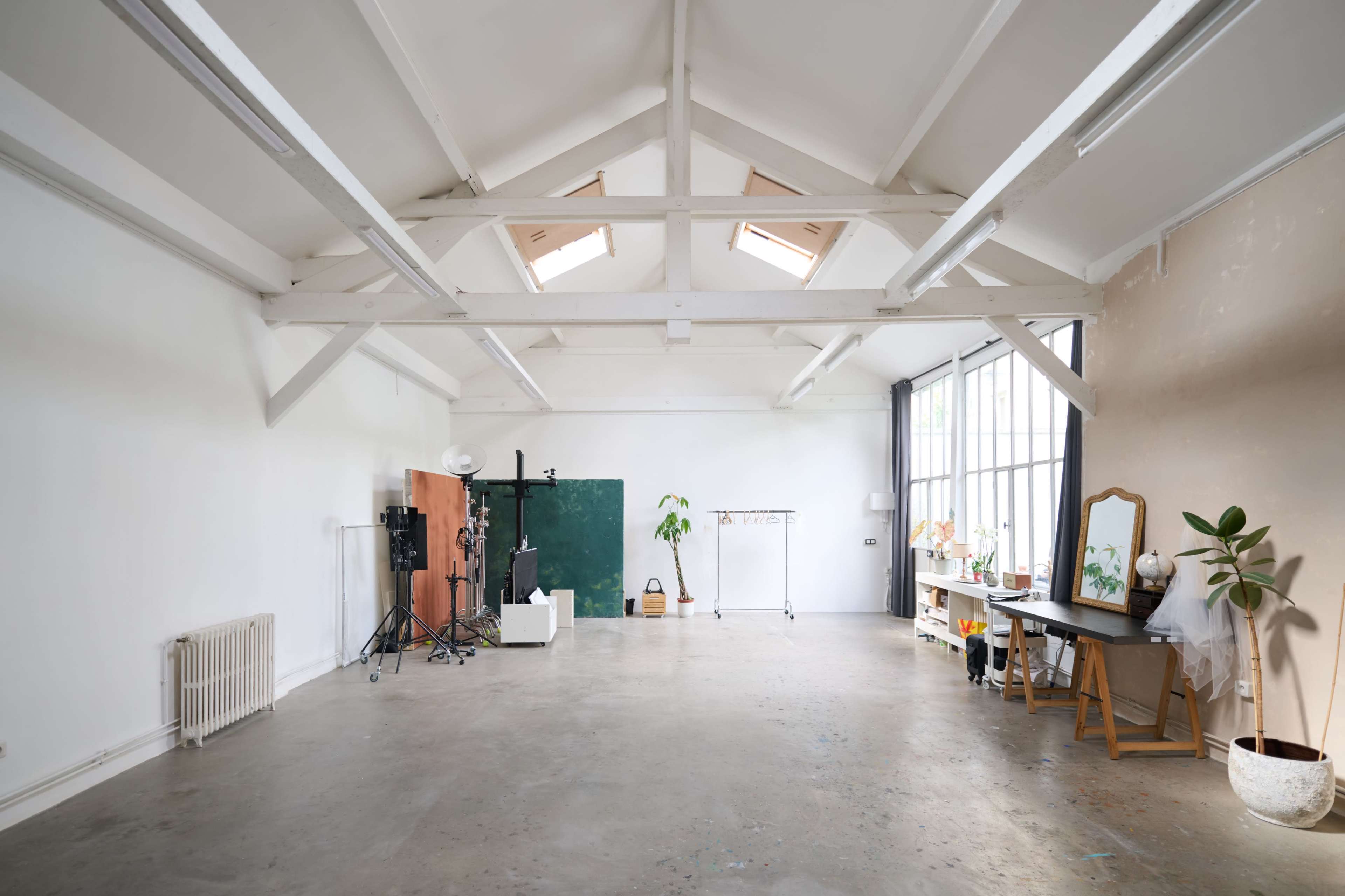 The image shows a spacious, empty studio with high ceilings, exposed wooden beams, and large windows, furnished with minimal items including a mirror, plant, and various equipment.