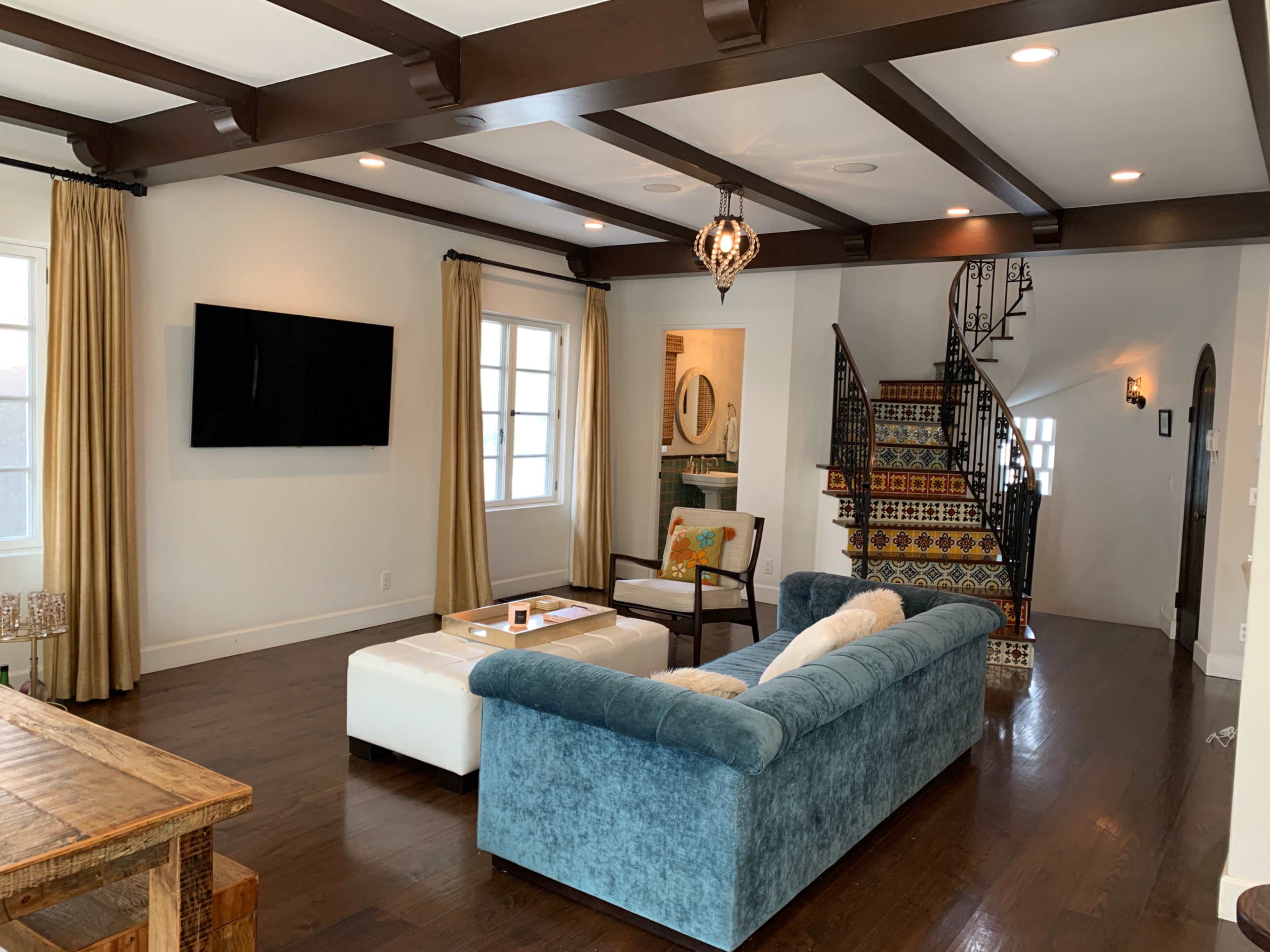 A living room with a blue sofa, a coffee table, and a staircase adorned with patterned tiles, all set in a space with wooden beams and bright windows.
