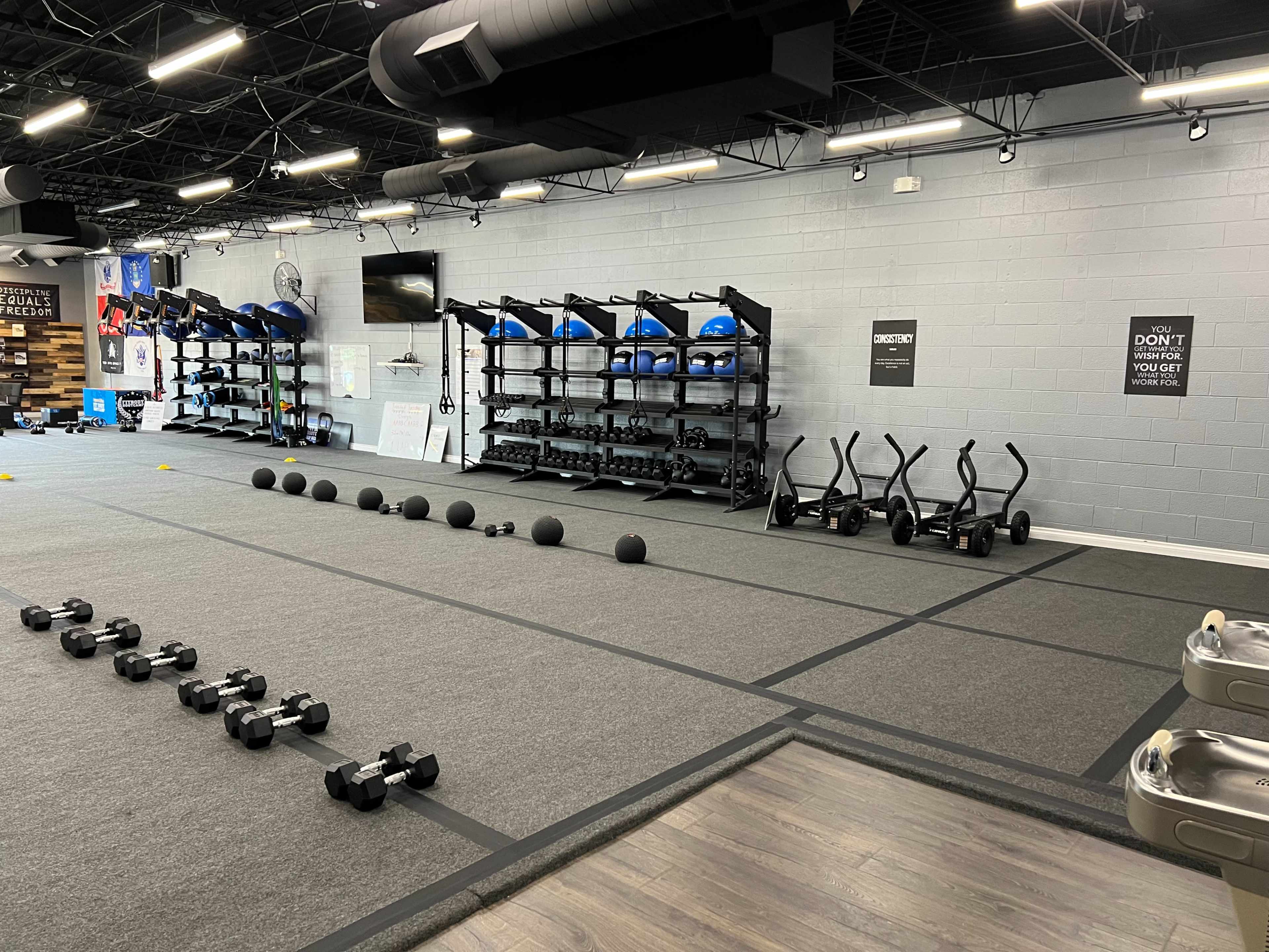 A fitness studio with various exercise equipment, including dumbbells, kettlebells, medicine balls, and stability balls organized on racks along the wall.