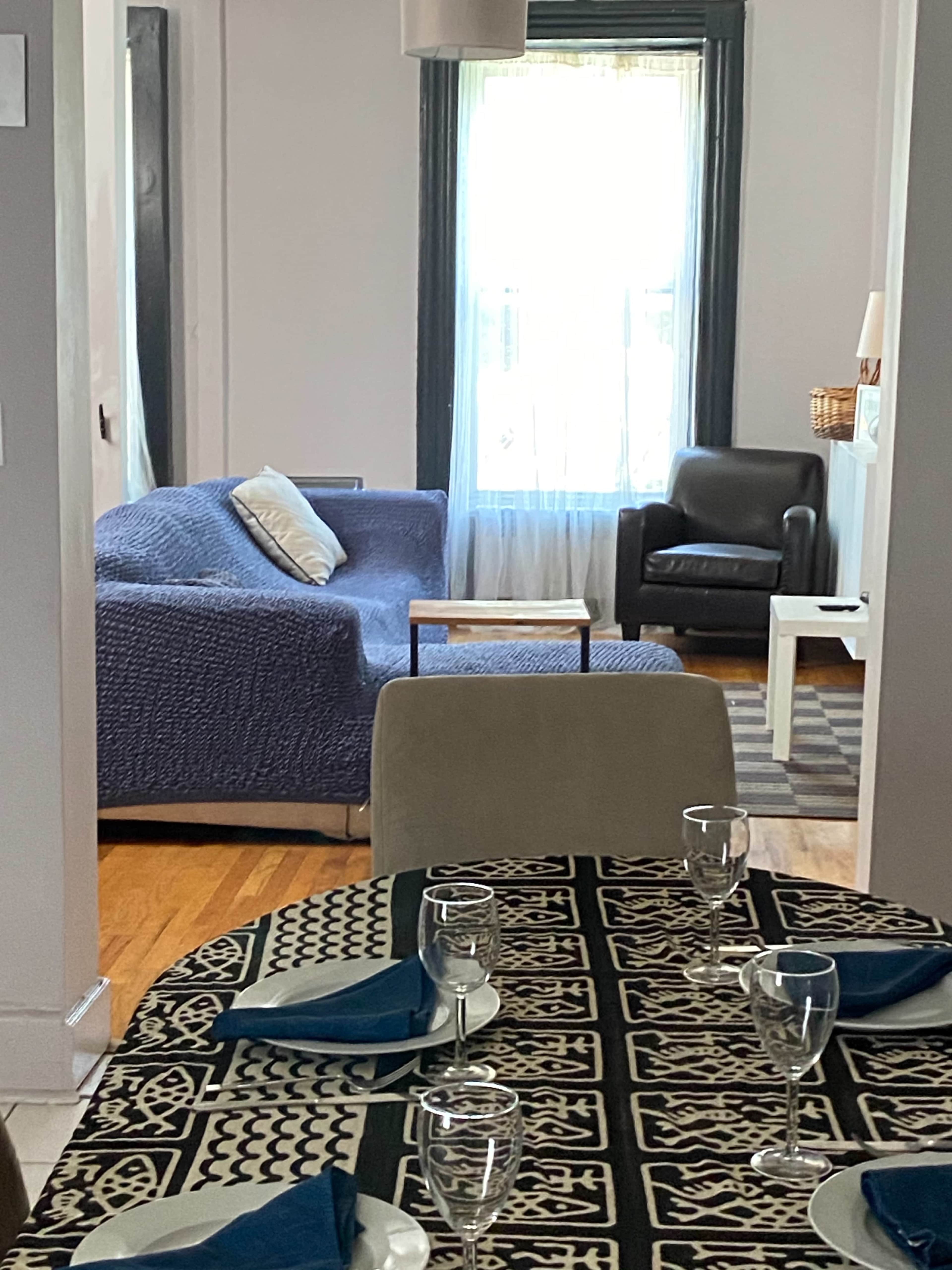 A dining table is set with glassware and blue napkins, leading into a living area featuring a blue sofa and a black chair beside a window.