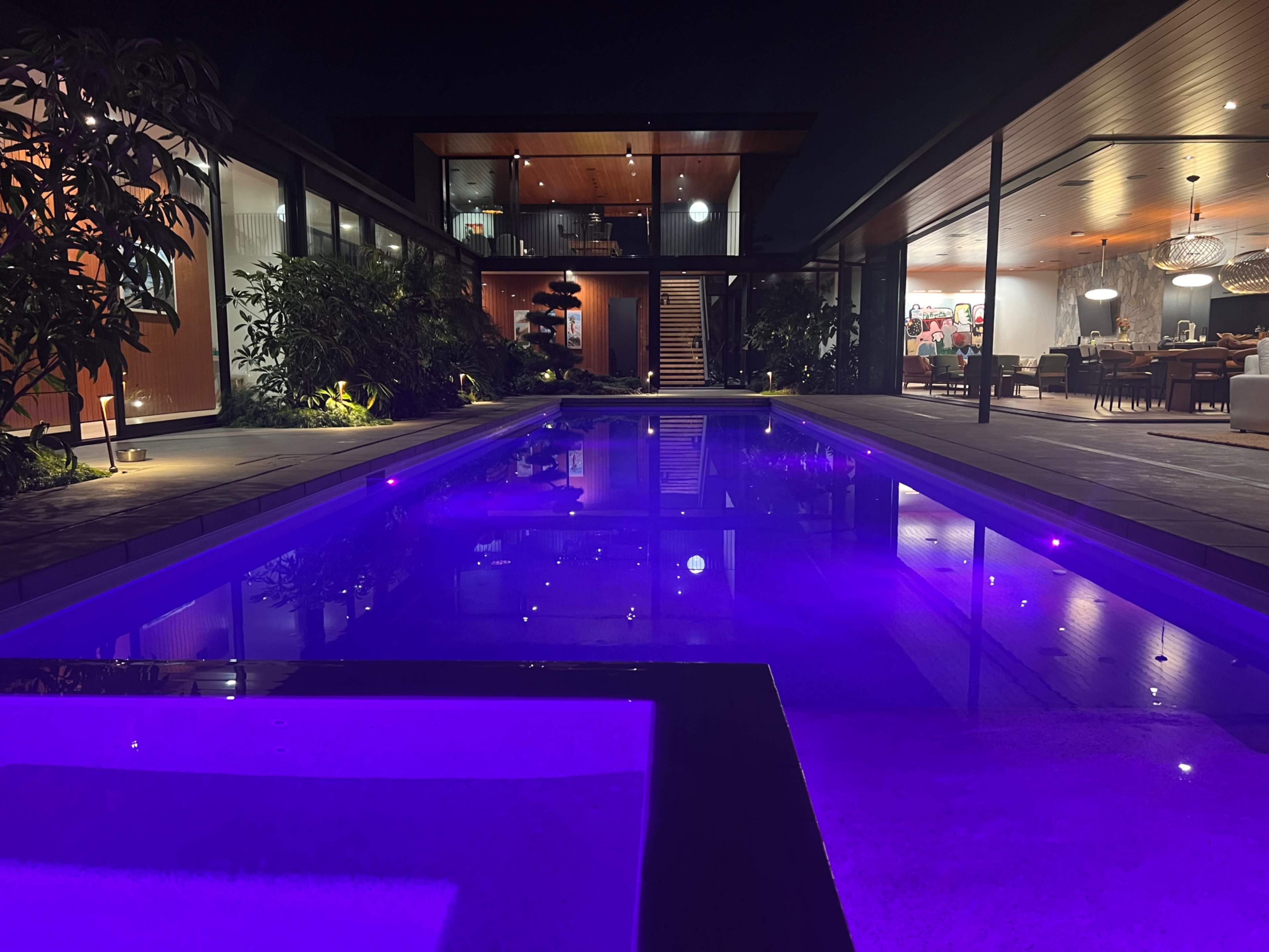 A modern home with a nighttime pool area illuminated by purple lights, surrounded by greenery and visible indoor spaces.