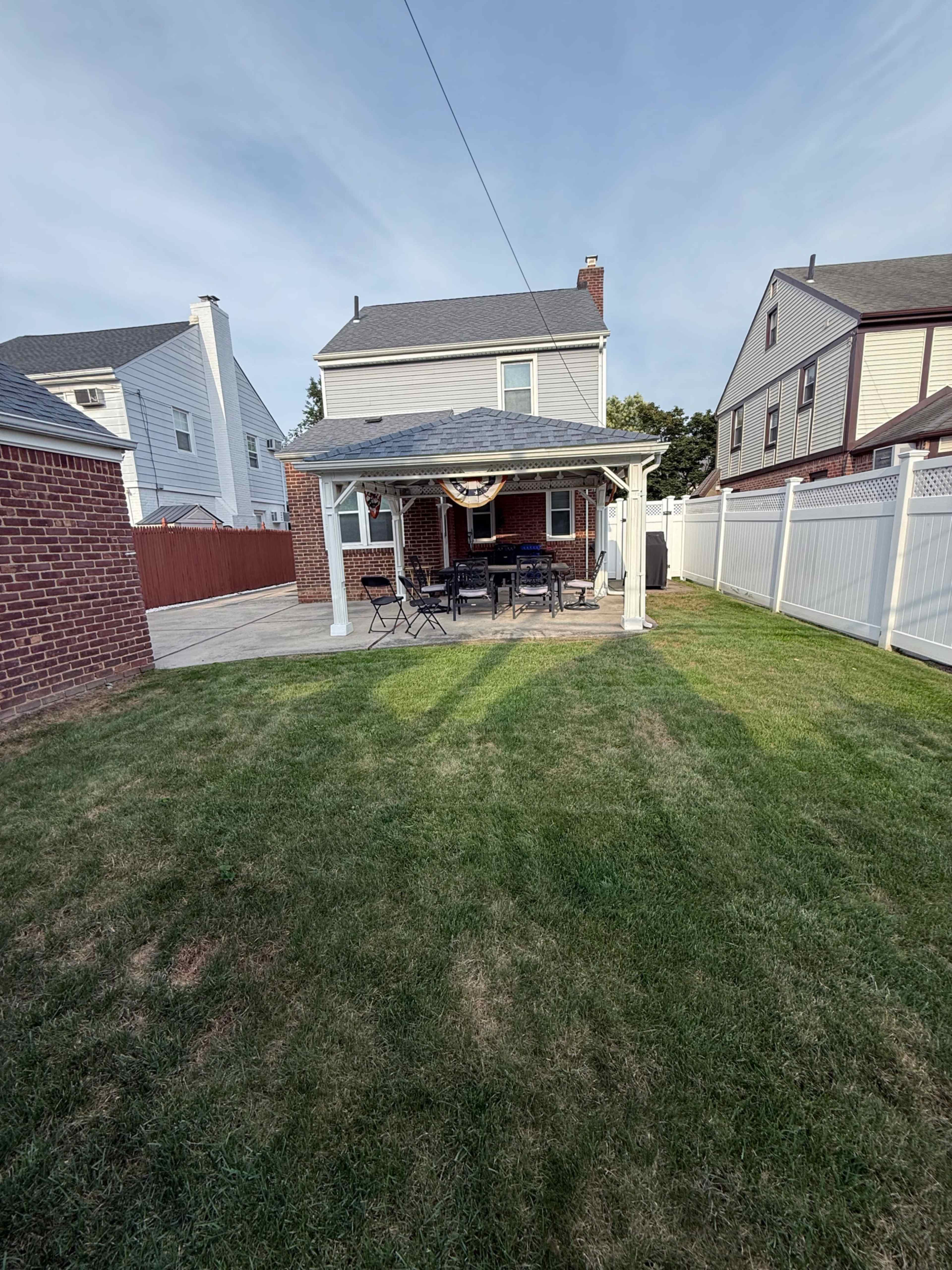 The image shows a backyard with a gazebo and a patio set, surrounded by a neatly trimmed lawn and residential homes.