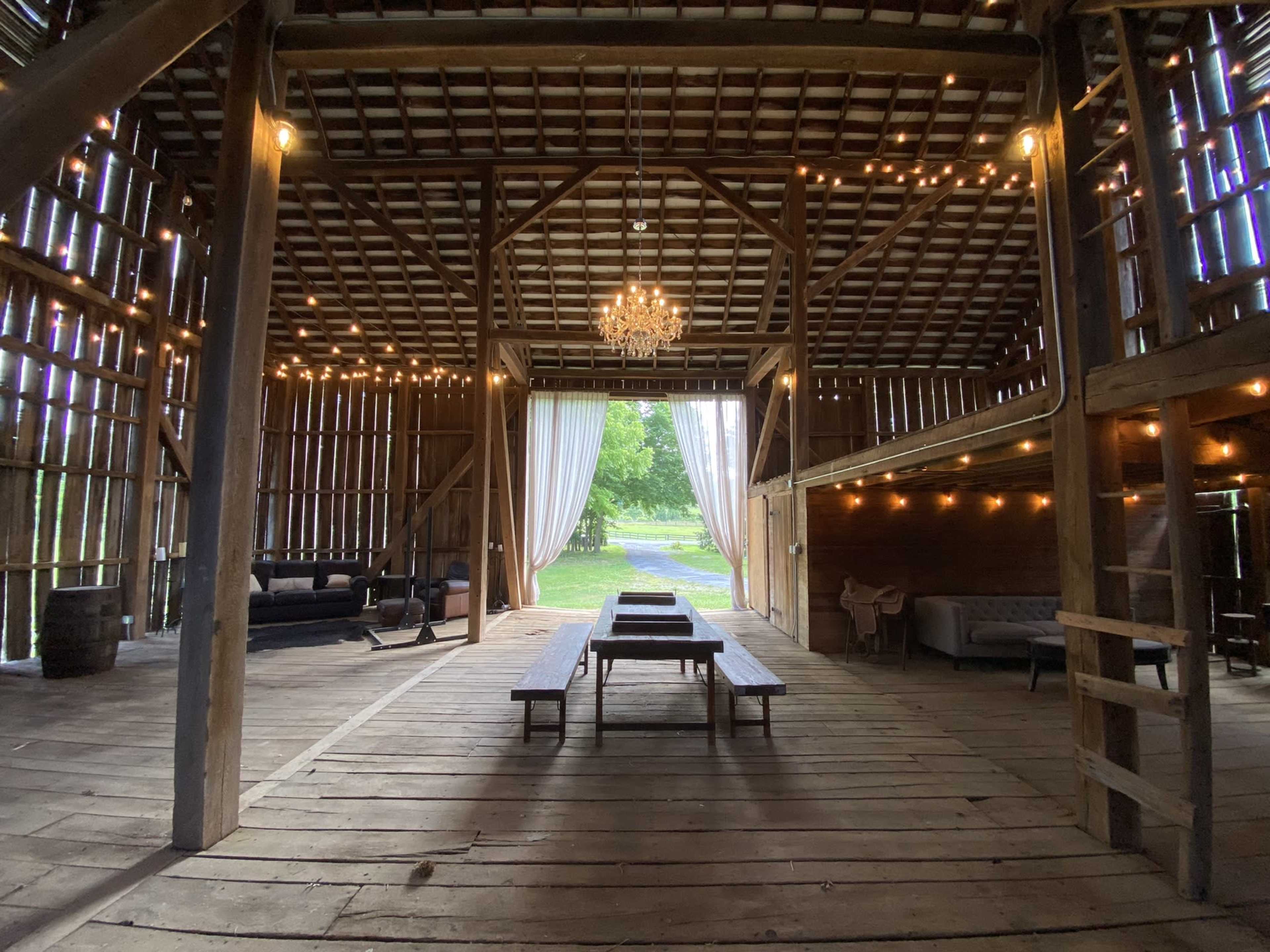 The interior of a rustic barn features wooden beams, string lights, and an elegant chandelier, with seating areas and large open doors leading to a landscaped view.