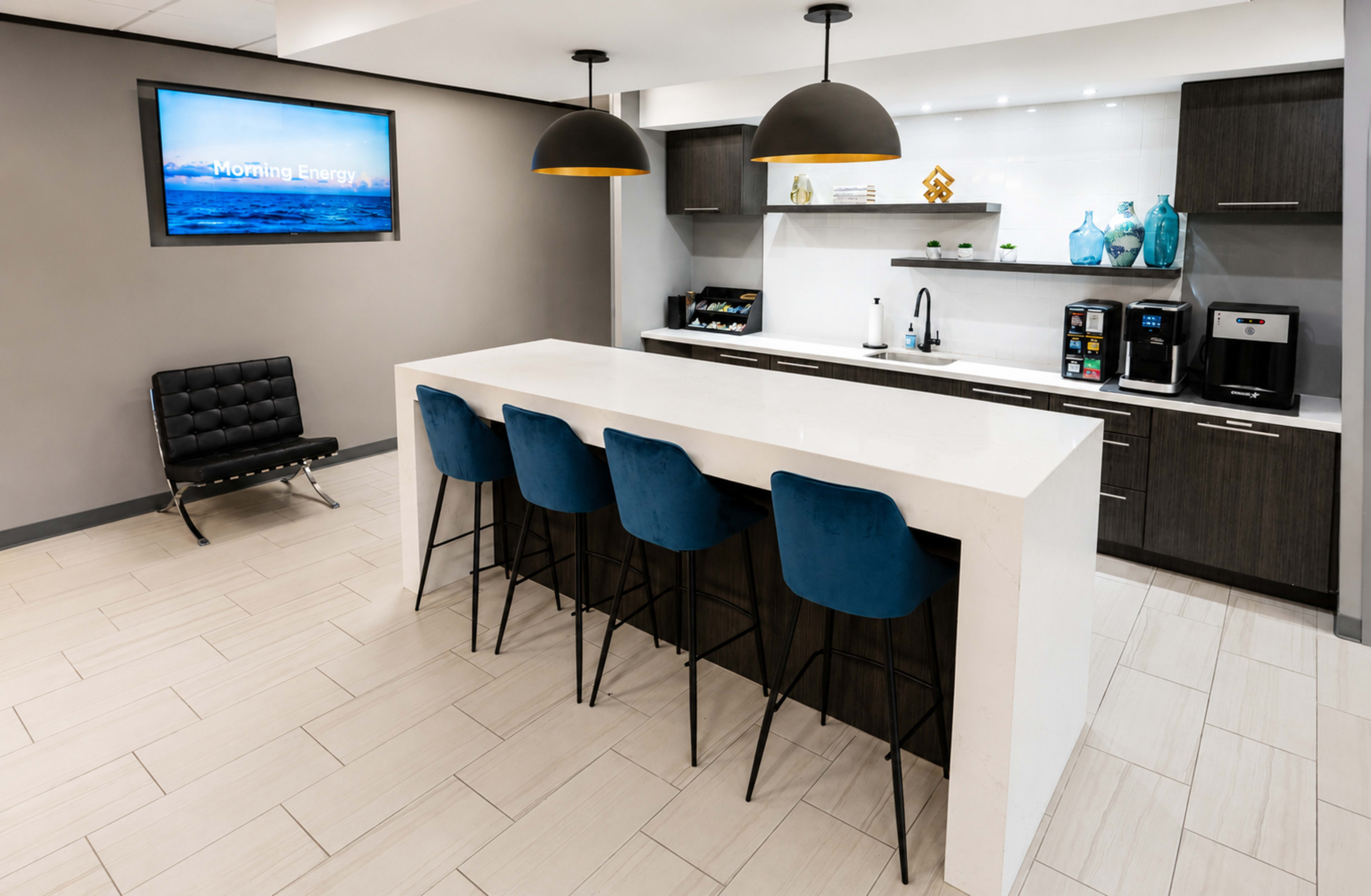 The image shows a modern kitchen space with a large white island, four blue bar stools, dark cabinetry, and a coffee station, complemented by pendant lighting and a wall-mounted screen displaying "Morning Energy."