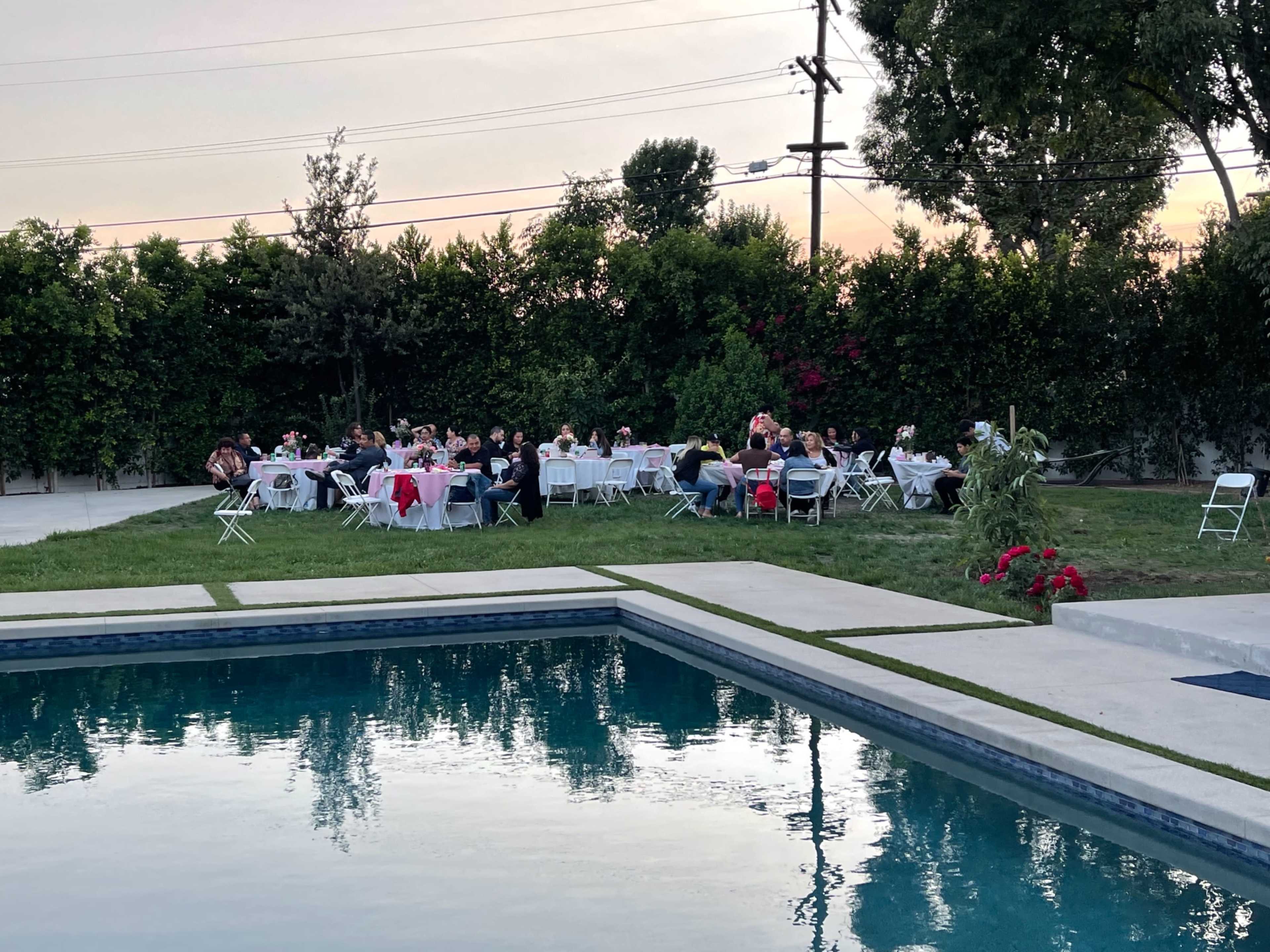 A group of people sits around tables set up for an outdoor event beside a swimming pool at dusk.