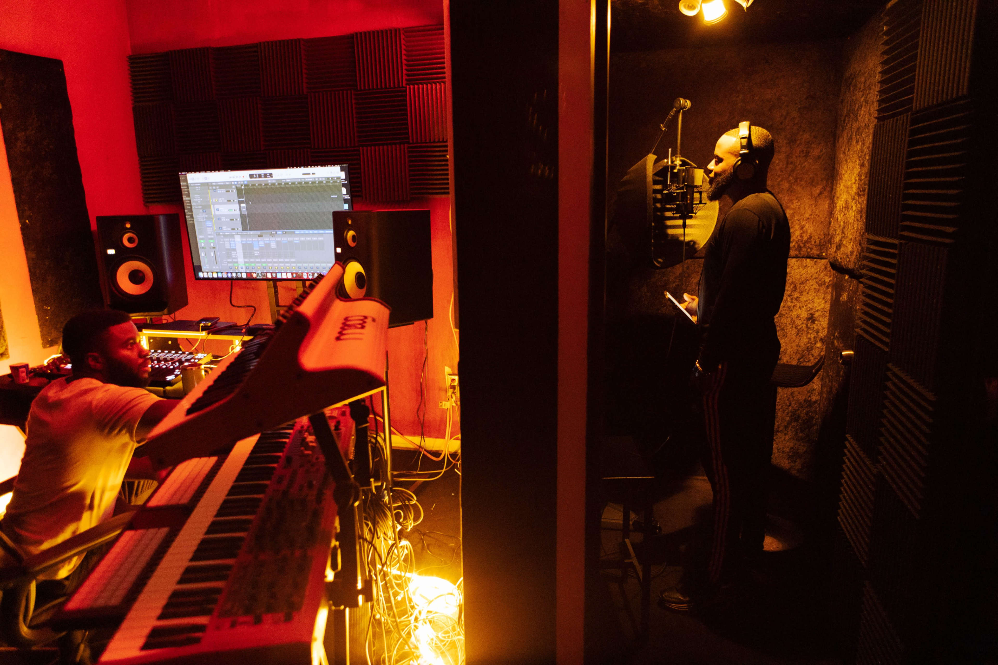 A musician stands in a soundproof booth recording vocals while another musician plays a synthesizer in a dimly lit studio.