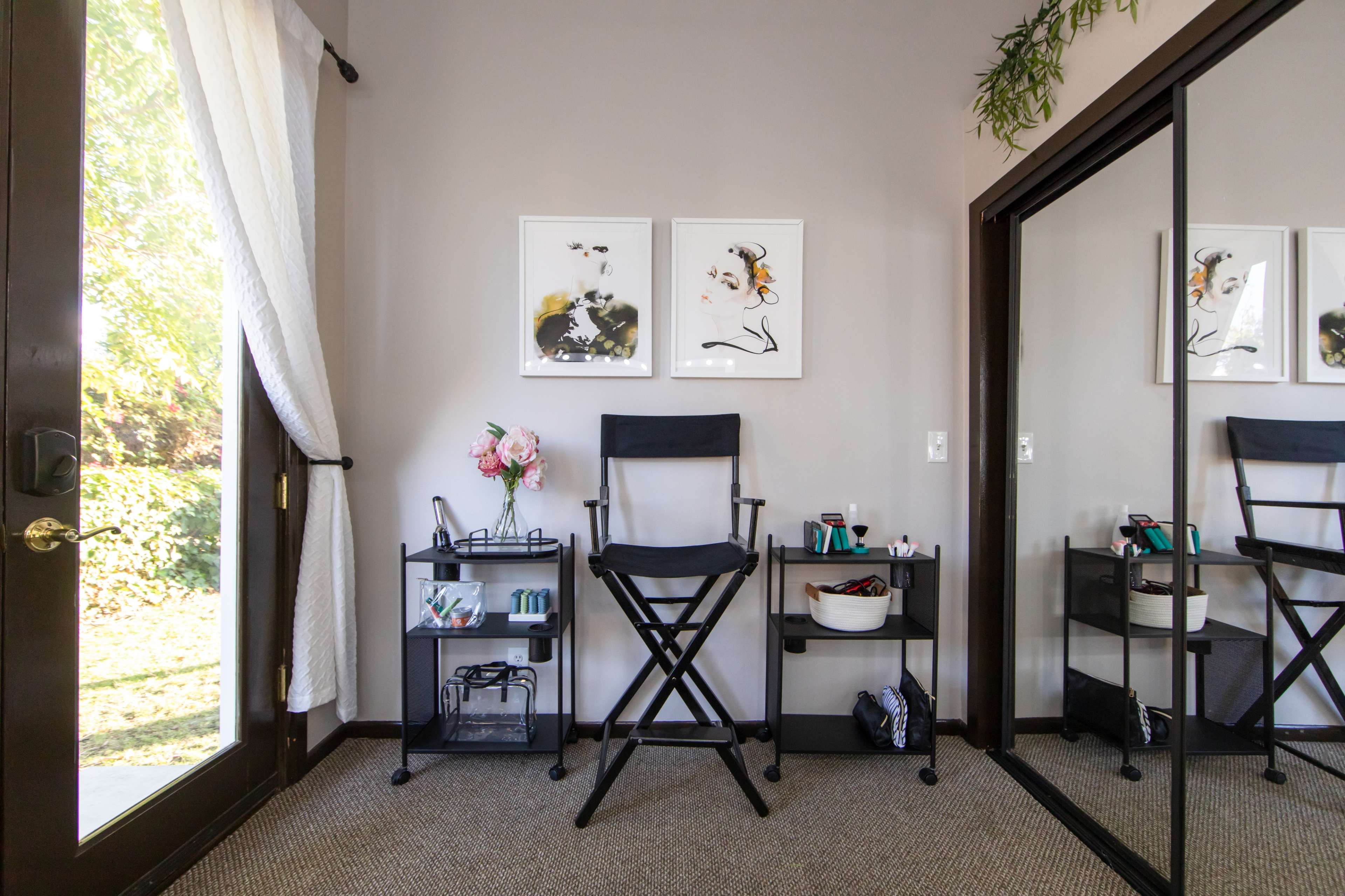 A makeup station with a black director's chair, two floral prints on the wall, and a mirror reflects shelves filled with cosmetics and a bouquet of flowers.