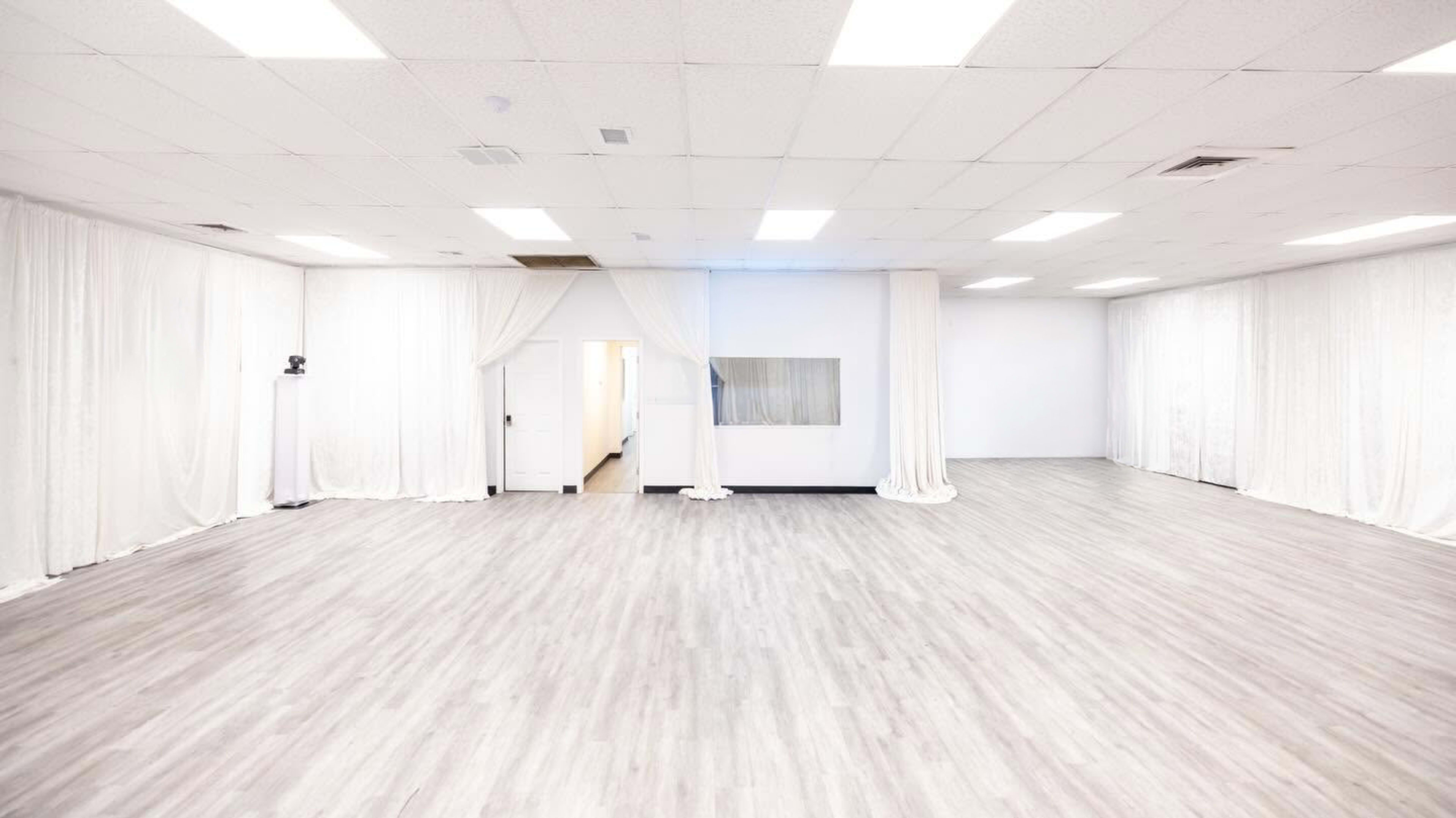 The image shows a spacious, well-lit room with light-colored flooring and white fabric drapes along the walls.