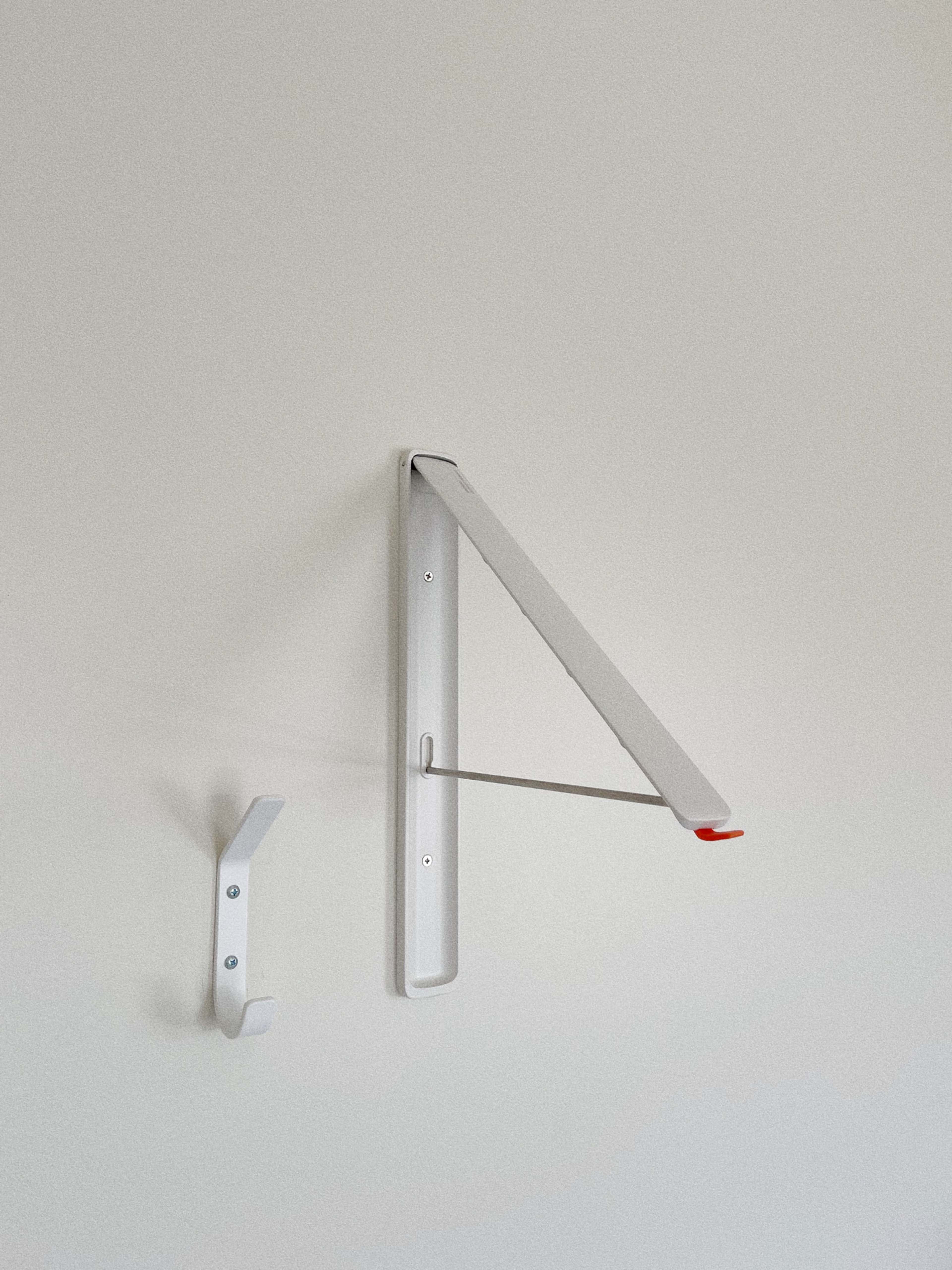The image features a minimalist wall-mounted coat rack and a folding shelf in a light-colored room.