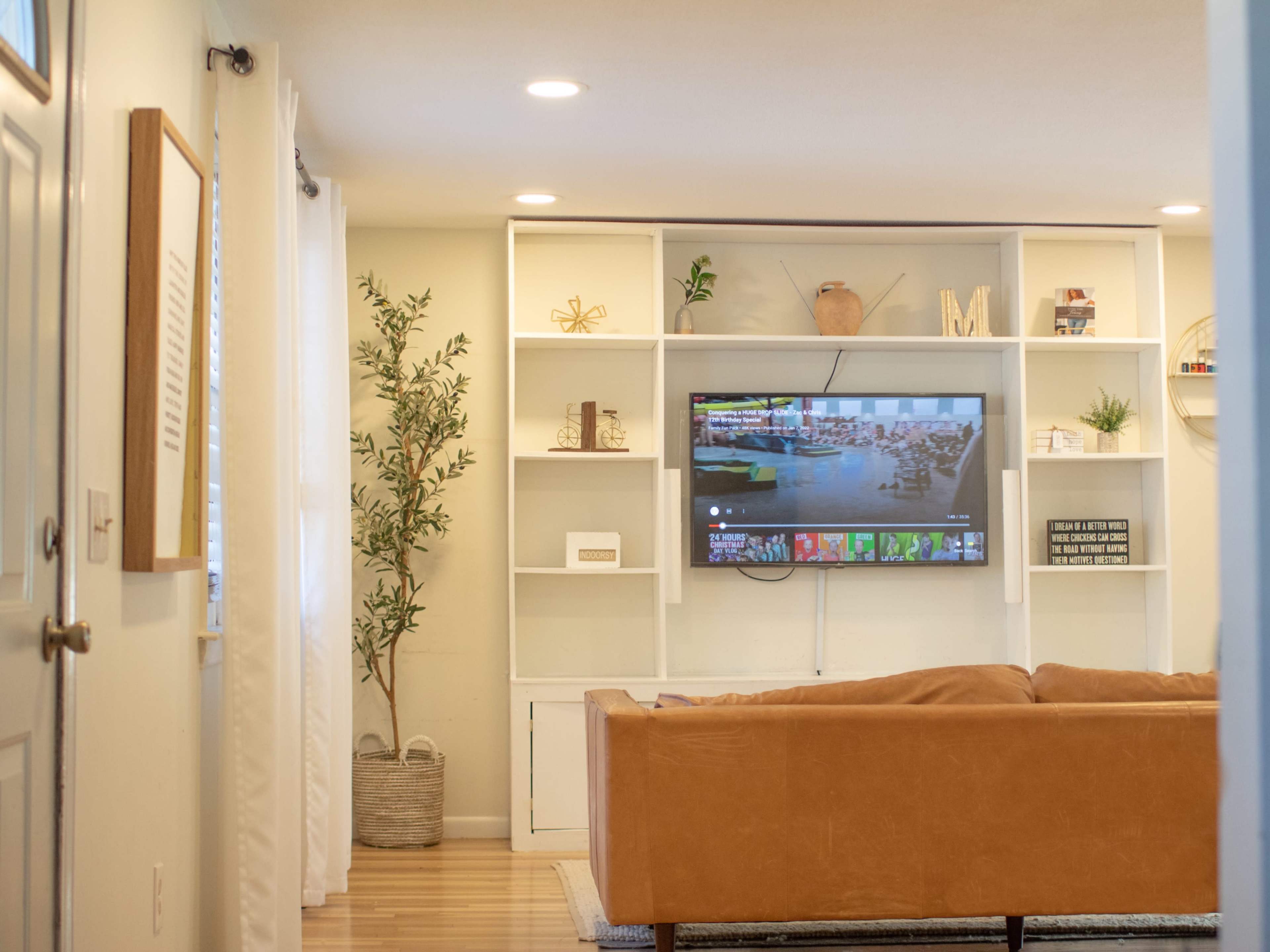A living room features a mounted television above a white shelving unit with decorative items, a tan sofa, and a large potted plant in the corner.