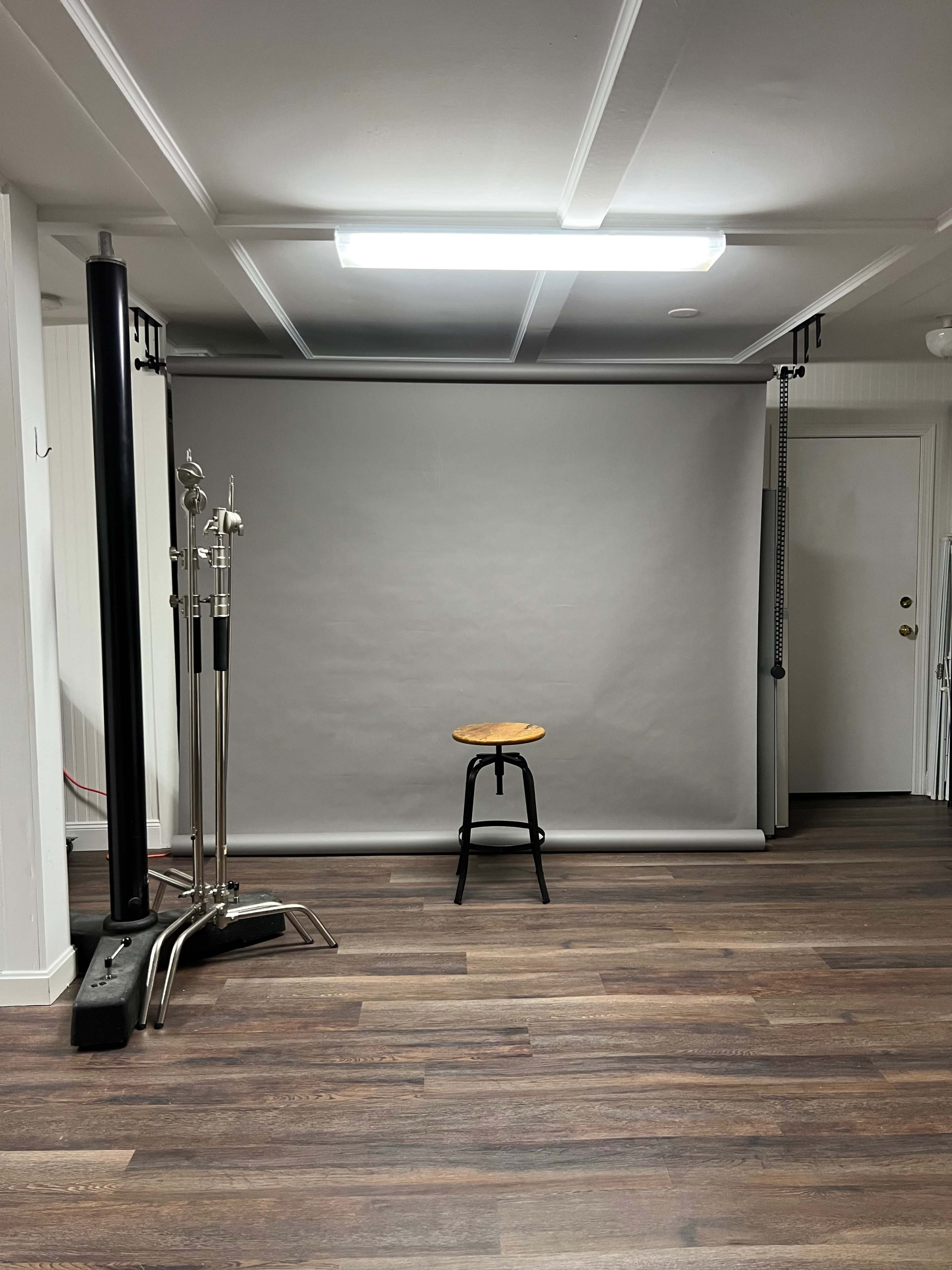 The image shows a photo studio with a gray backdrop, a wooden stool in the center, and studio lights on either side.