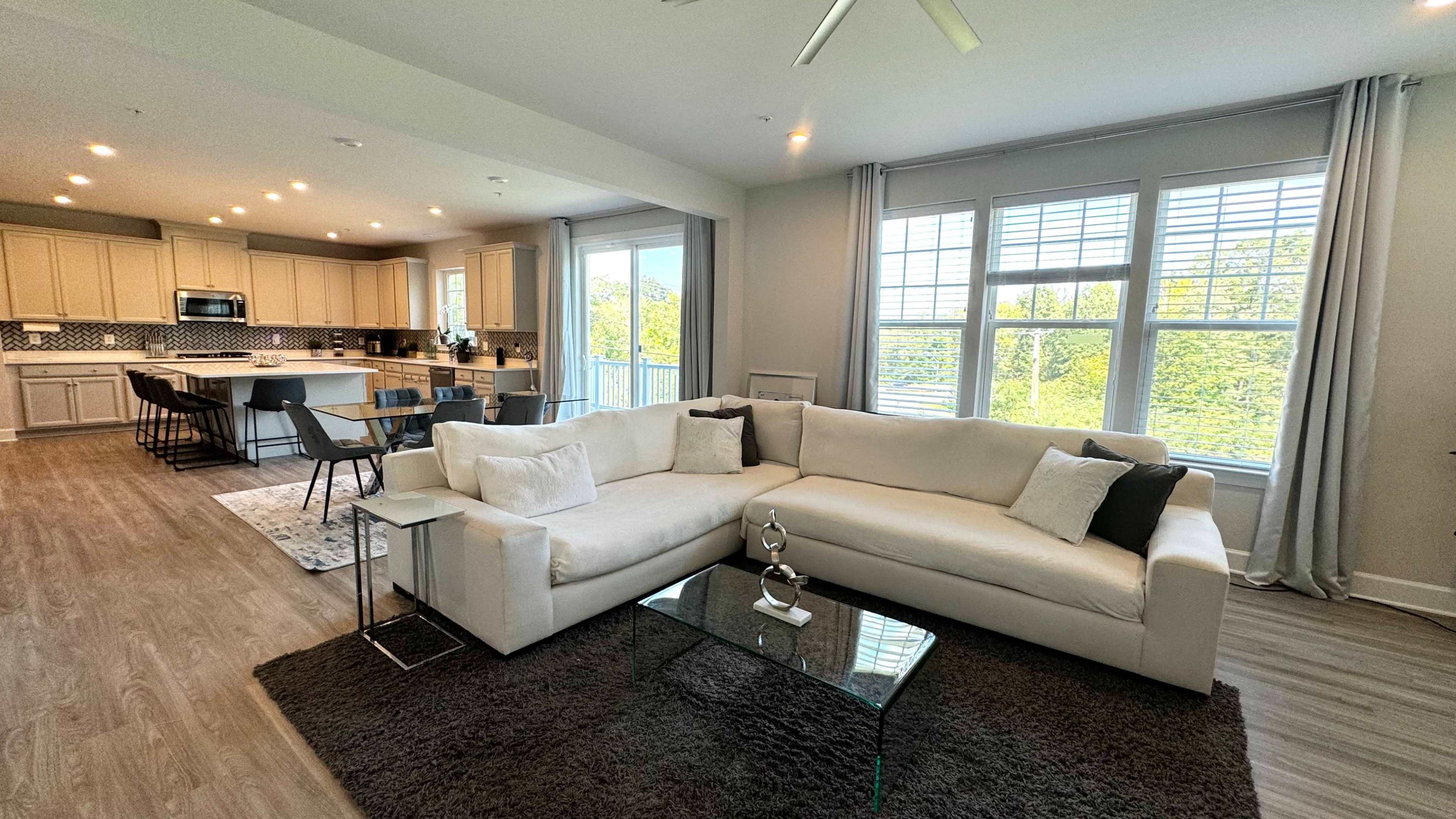 A modern living room with a white sectional sofa, a glass coffee table, and large windows allowing natural light, adjacent to a kitchen area with beige cabinetry and contrast tile.