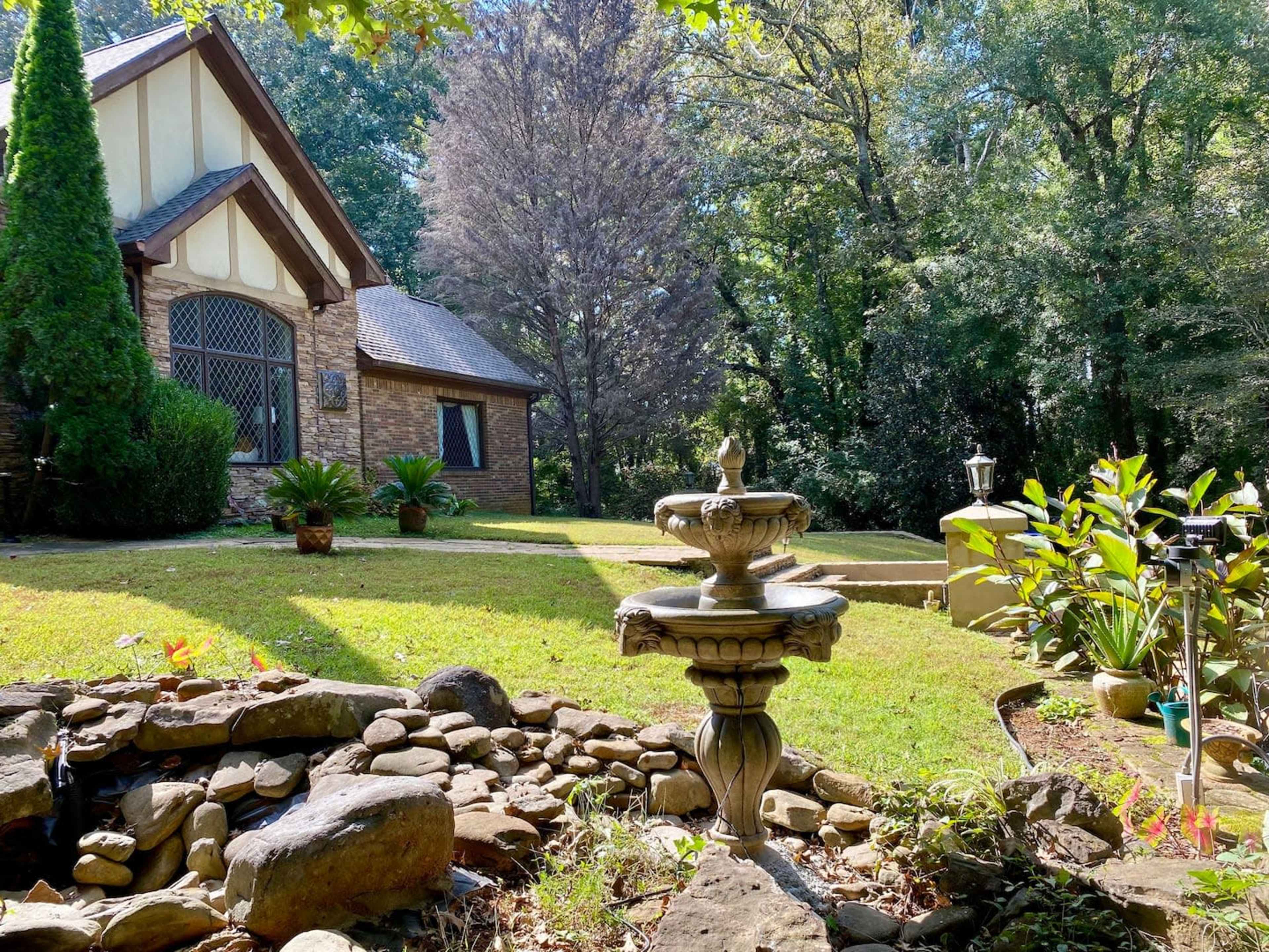 A stone fountain stands in a garden in front of a house surrounded by trees and landscaped plants.