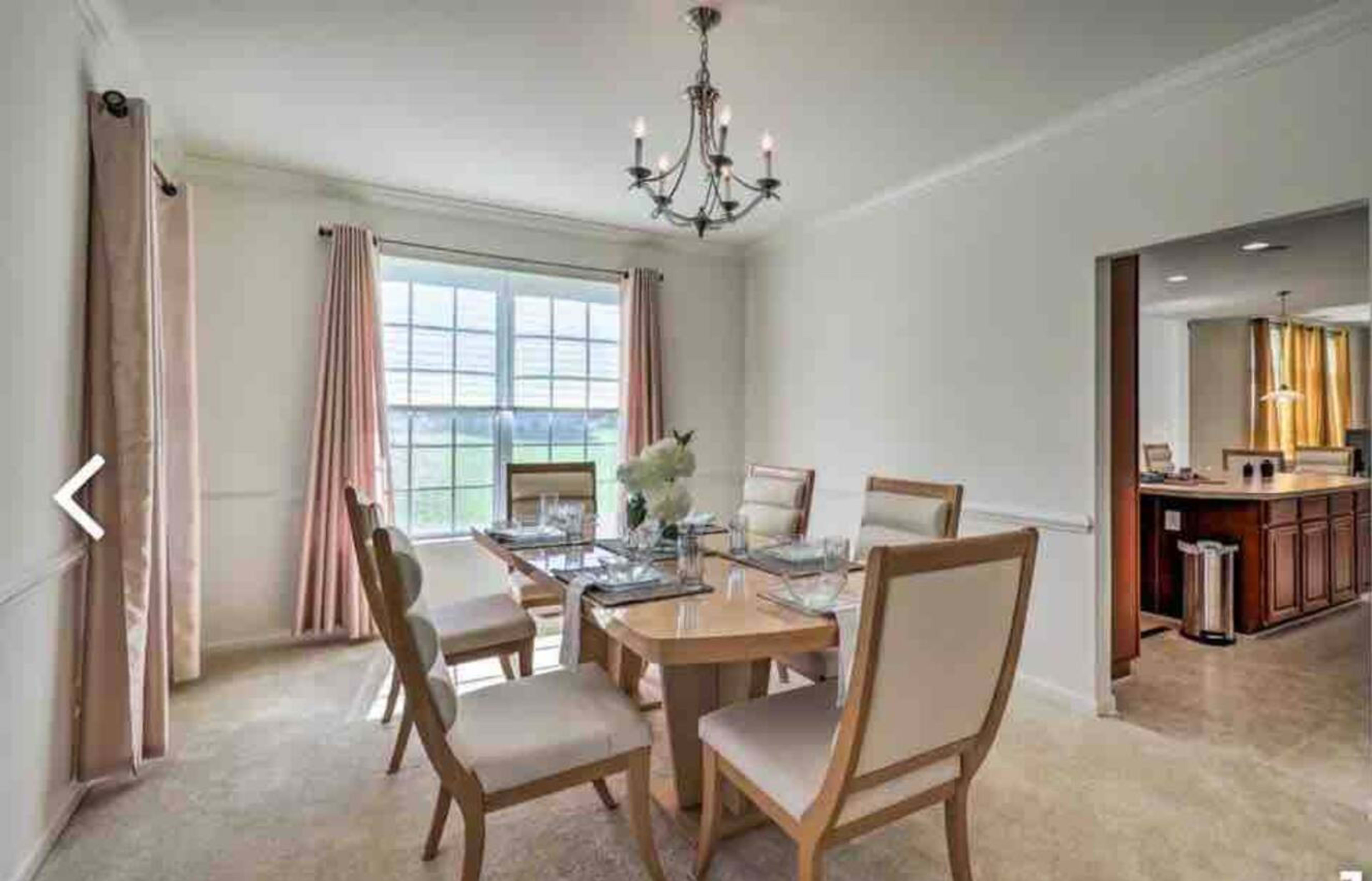 A dining room features a round table set for six, with a chandelier above and a large window letting in natural light.