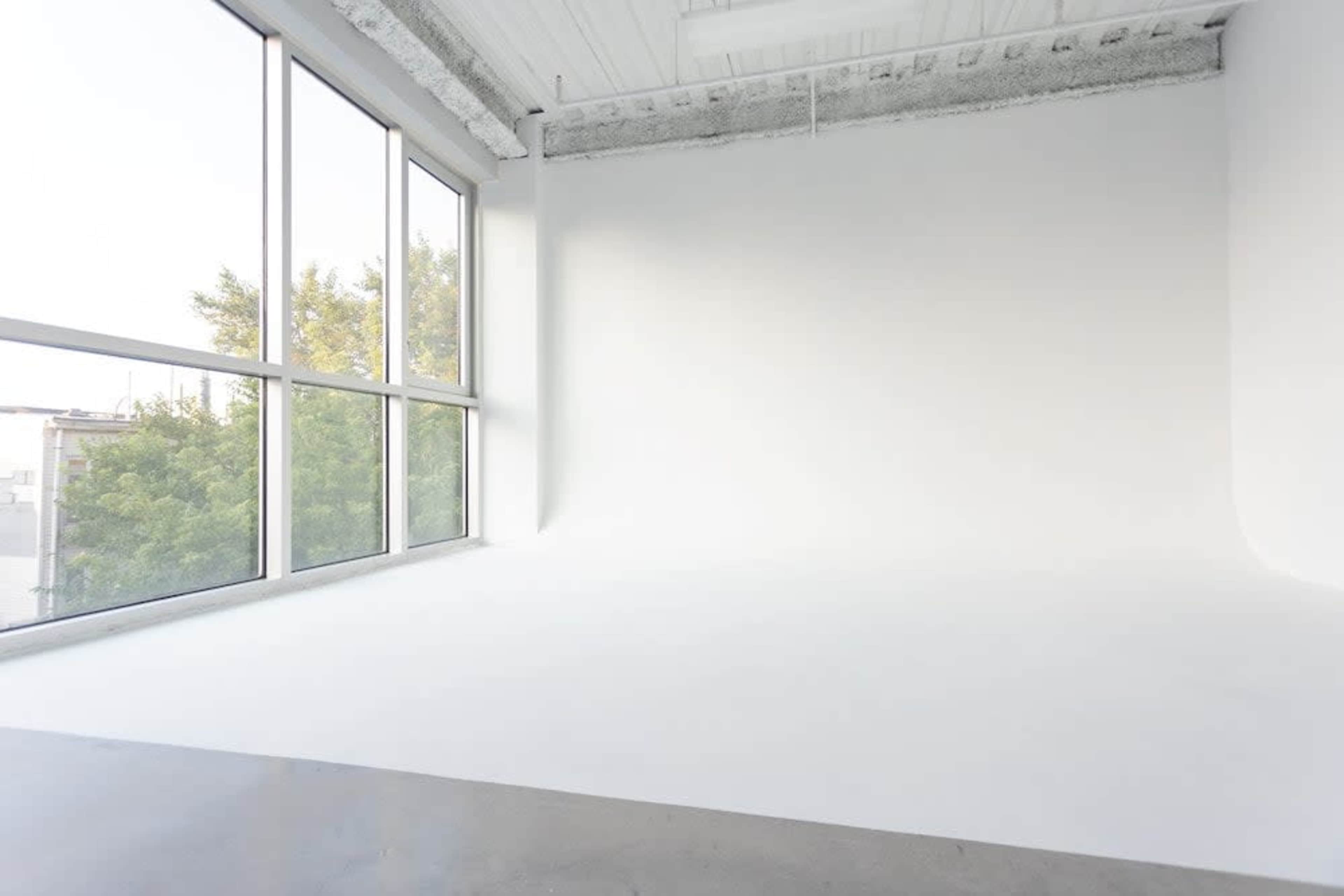 A spacious, empty room with large windows letting in natural light and a seamless white backdrop.
