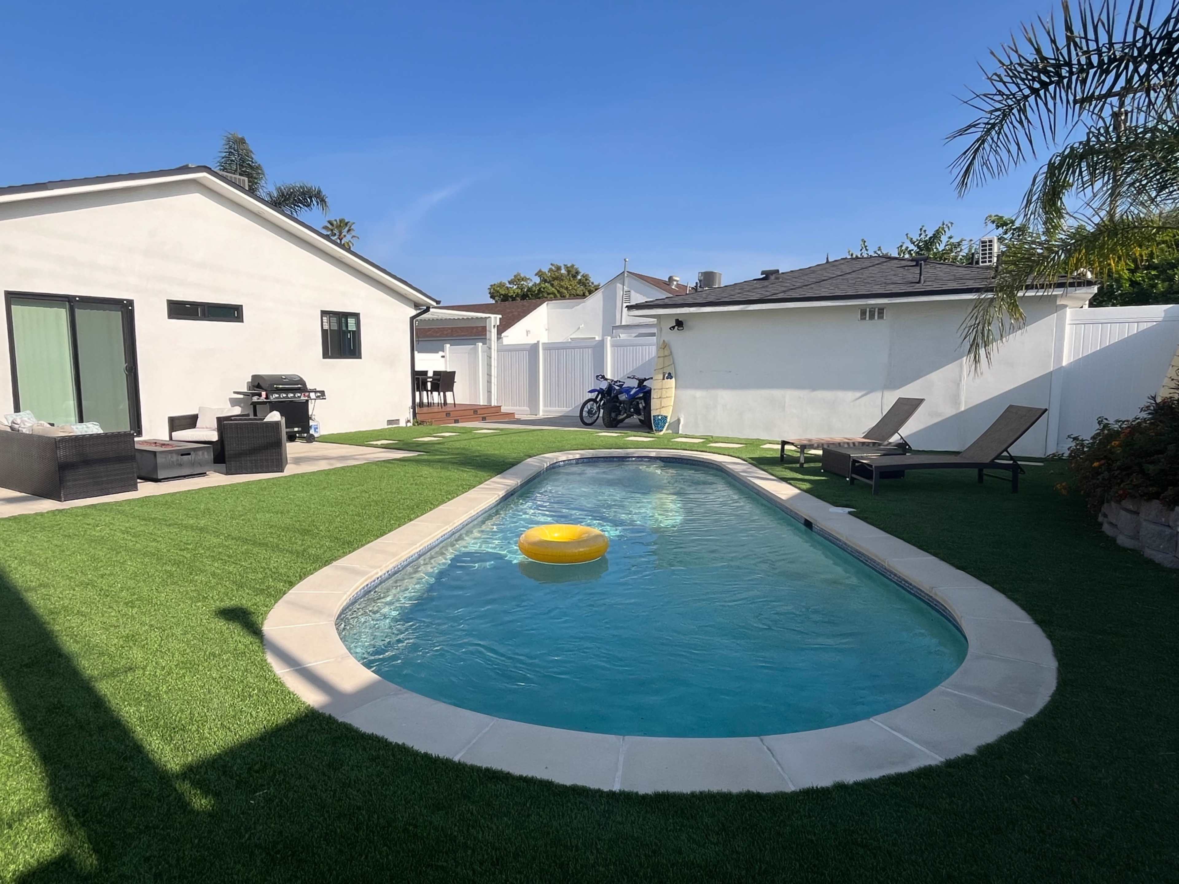 A brightly lit backyard features a swimming pool with a yellow float, surrounded by green grass and lounge chairs.