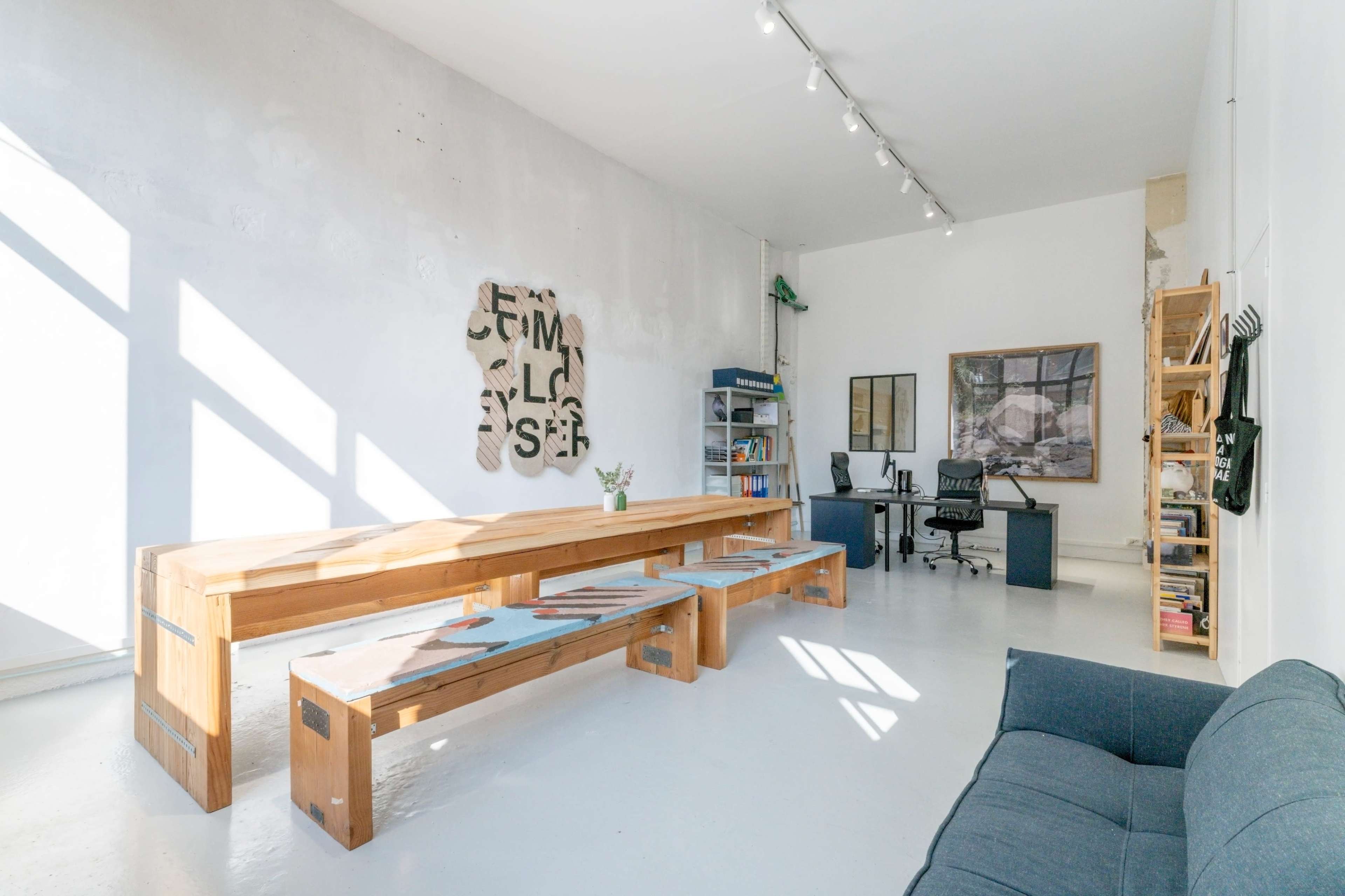 A minimalist office space featuring a large wooden table, a sofa, a desk with a chair, and a wall with abstract art.
