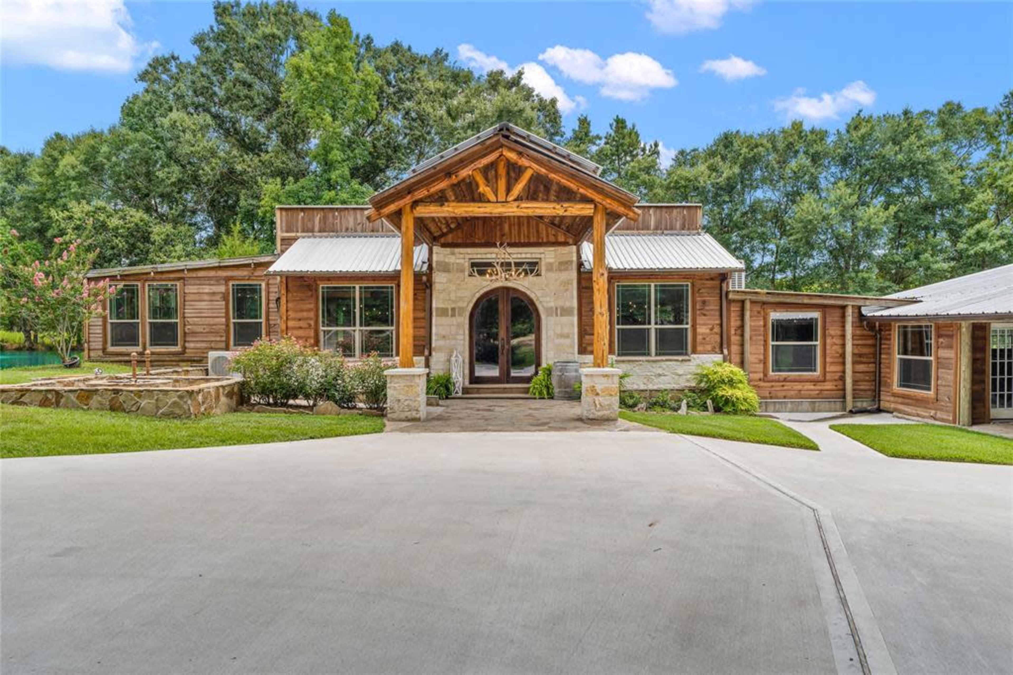 A large, wooden house with a prominent front porch and stone accents is surrounded by lush greenery.