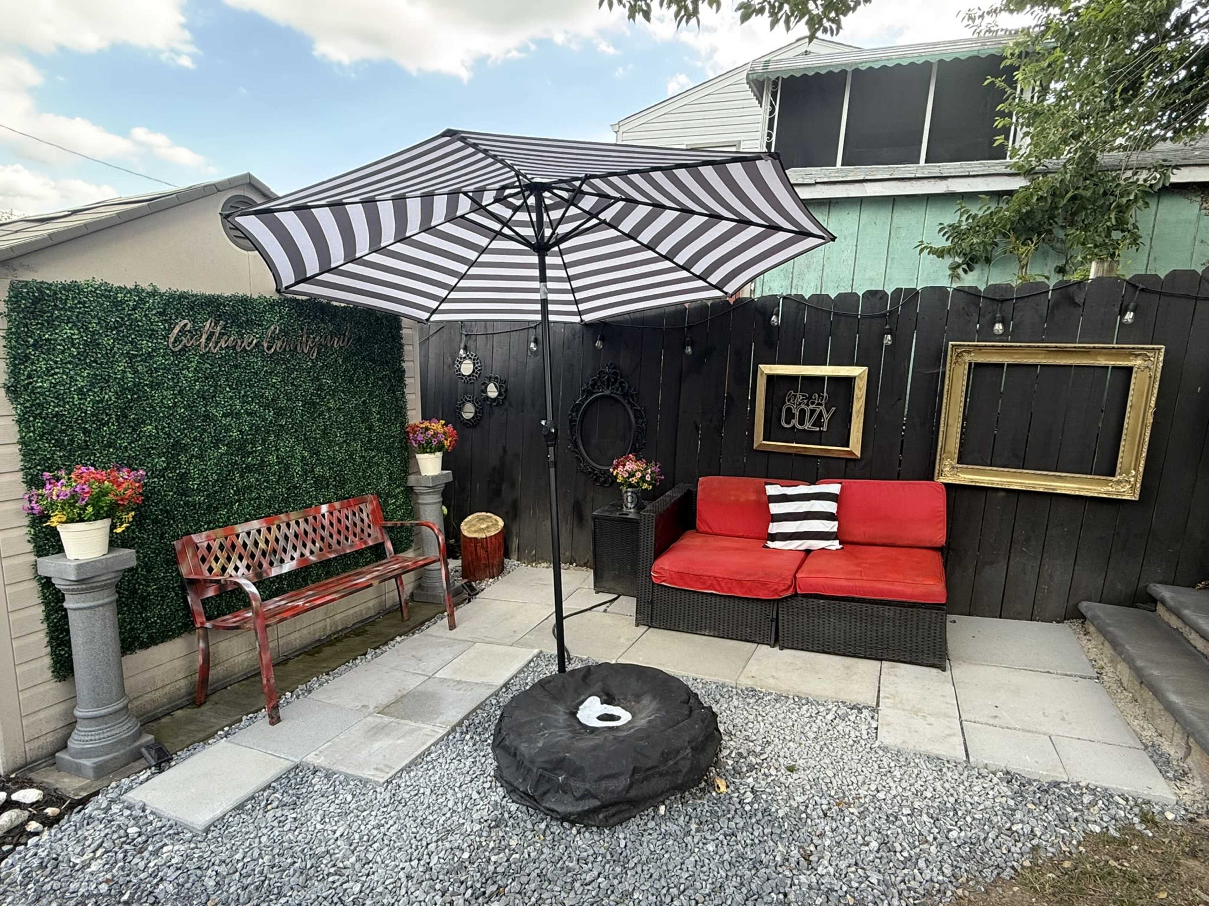 The image shows a small outdoor seating area featuring a red bench, a red couch with black and white cushions, a striped umbrella, and decorative planters on gravel flooring.