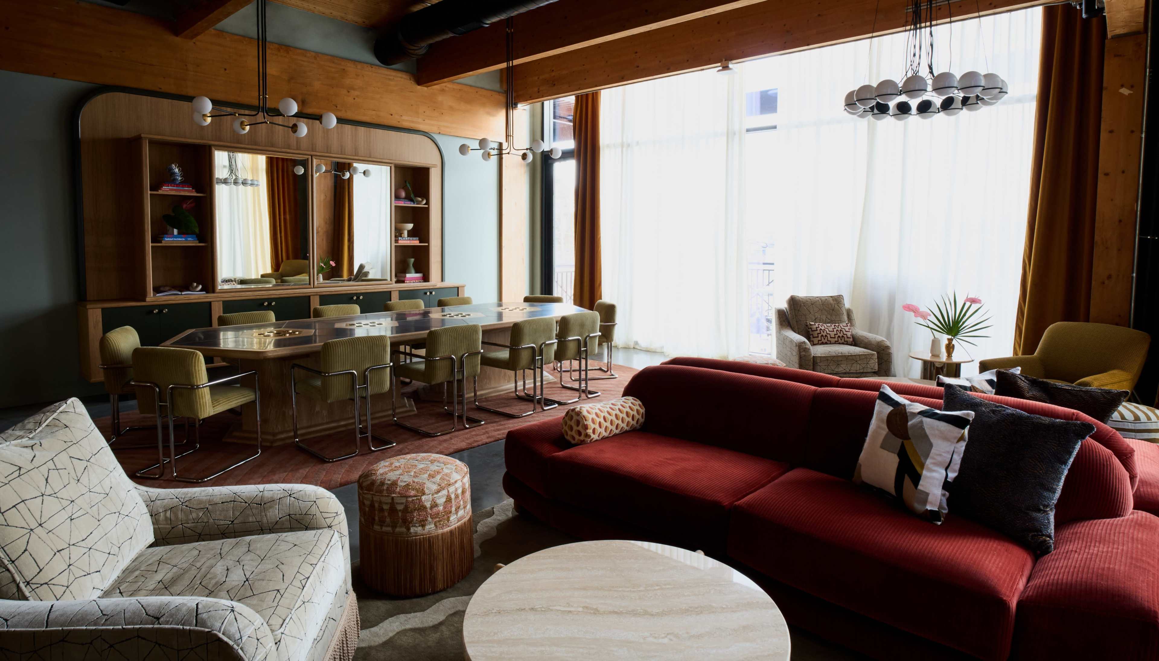 A modern living space featuring a red sofa, a round coffee table, and a dining area with green chairs and a large window allowing natural light to enter.