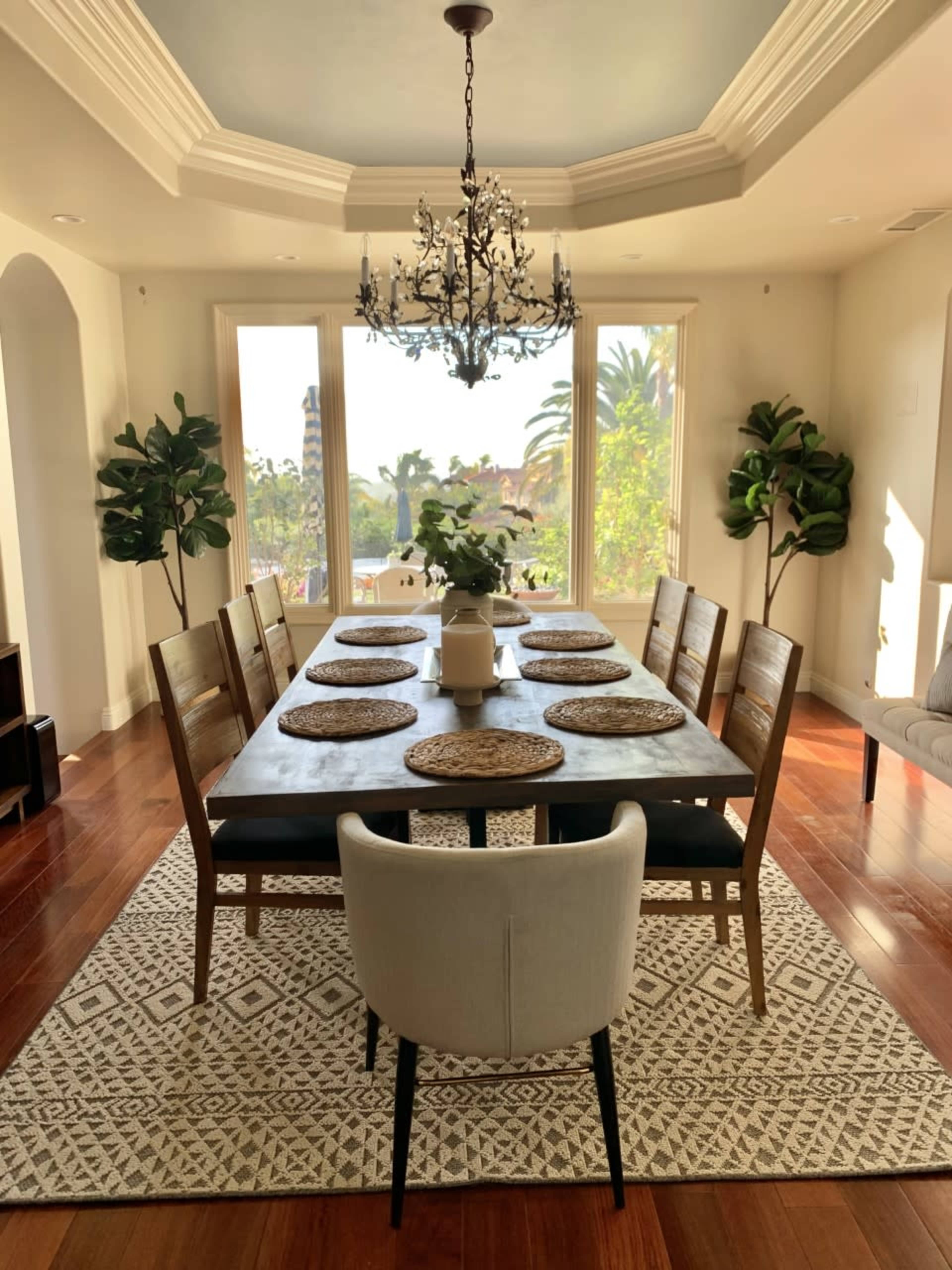 A spacious dining room features a long wooden table set with placemats, surrounded by chairs, under a chandelier with large windows showcasing greenery outside.